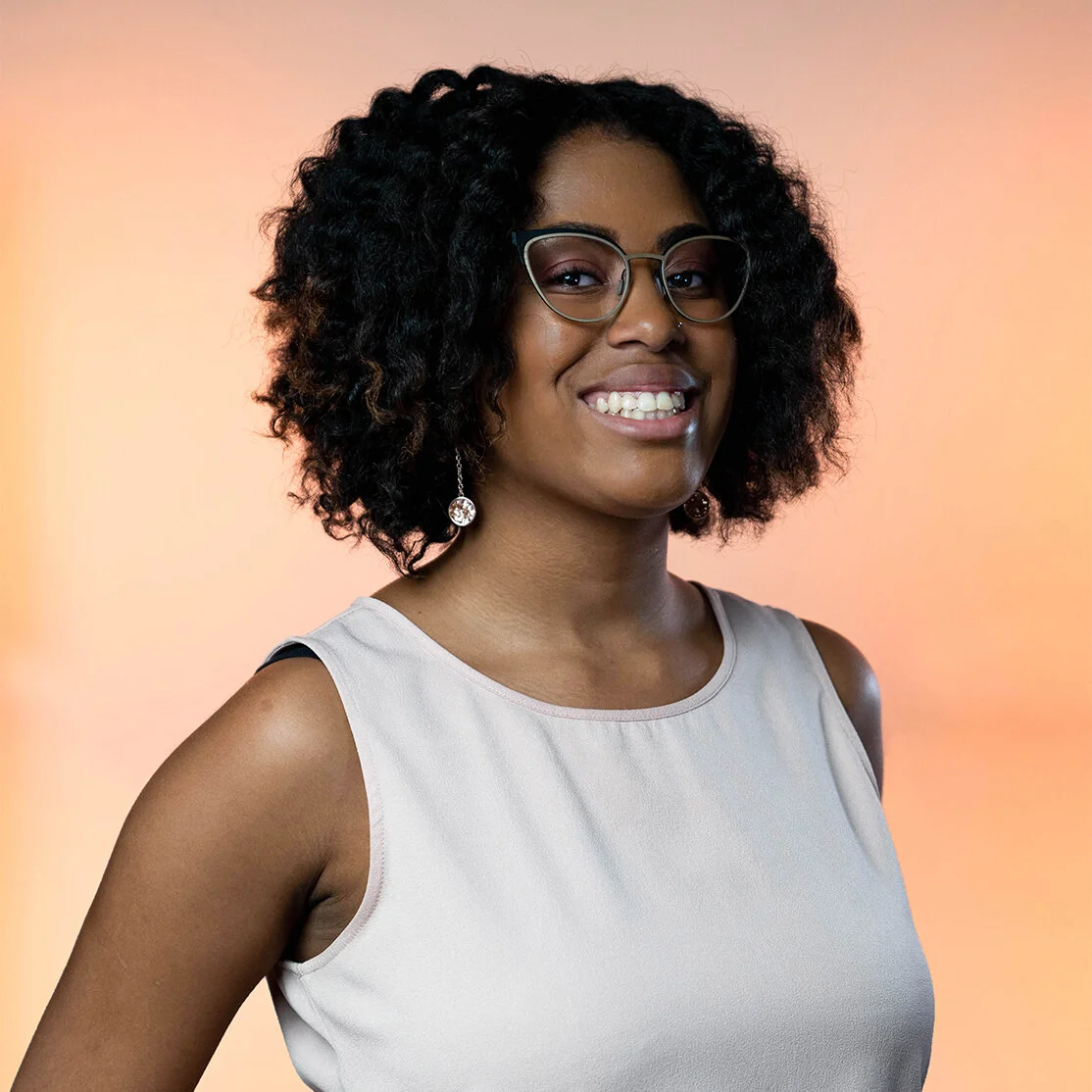A Black woman with shoulder length brown hair is smiling wearing glasses and a white tank top standing against a light orange background