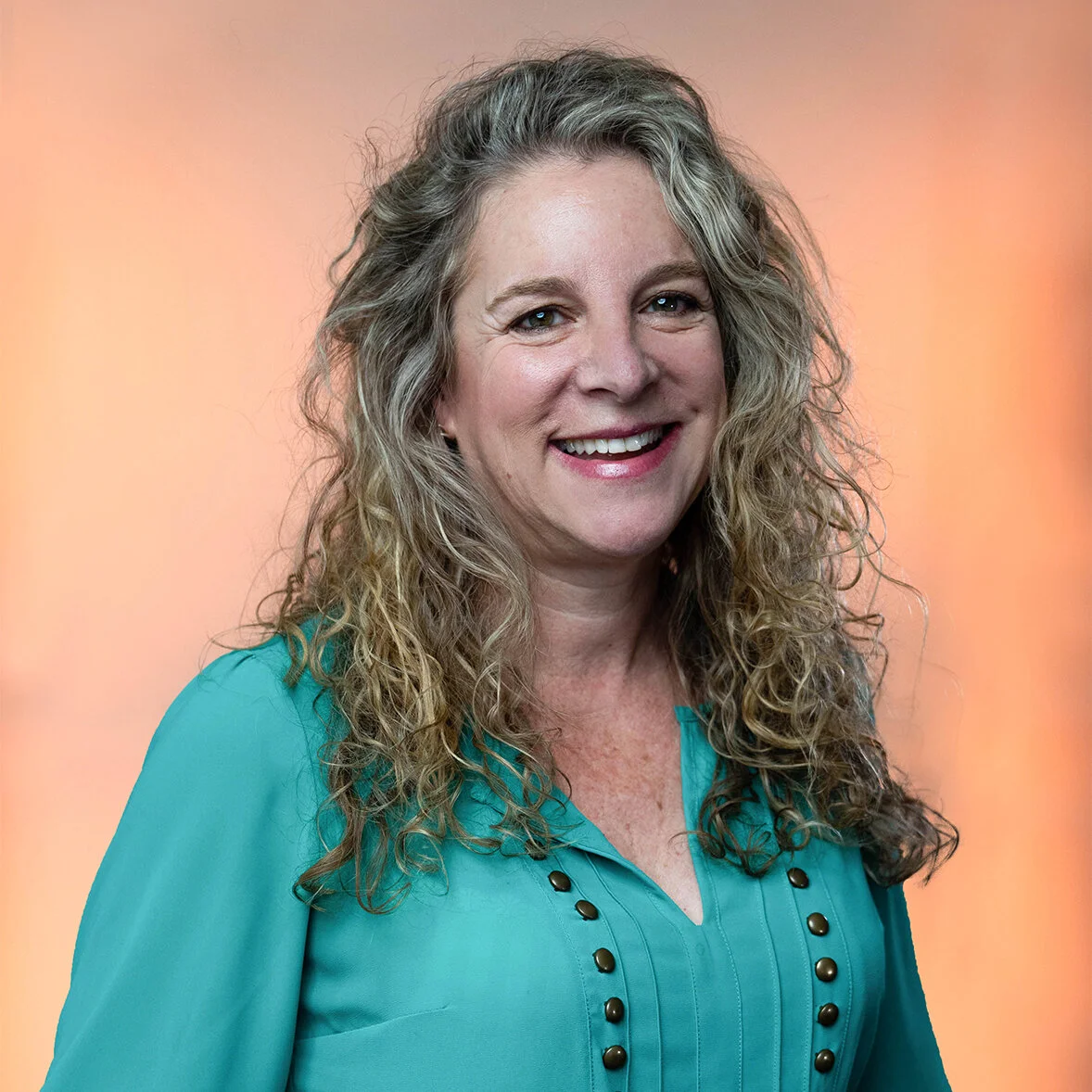 A white woman with long curly blonde hair is smiling wearing a teal blouse with buttons and is standing against a light orange background