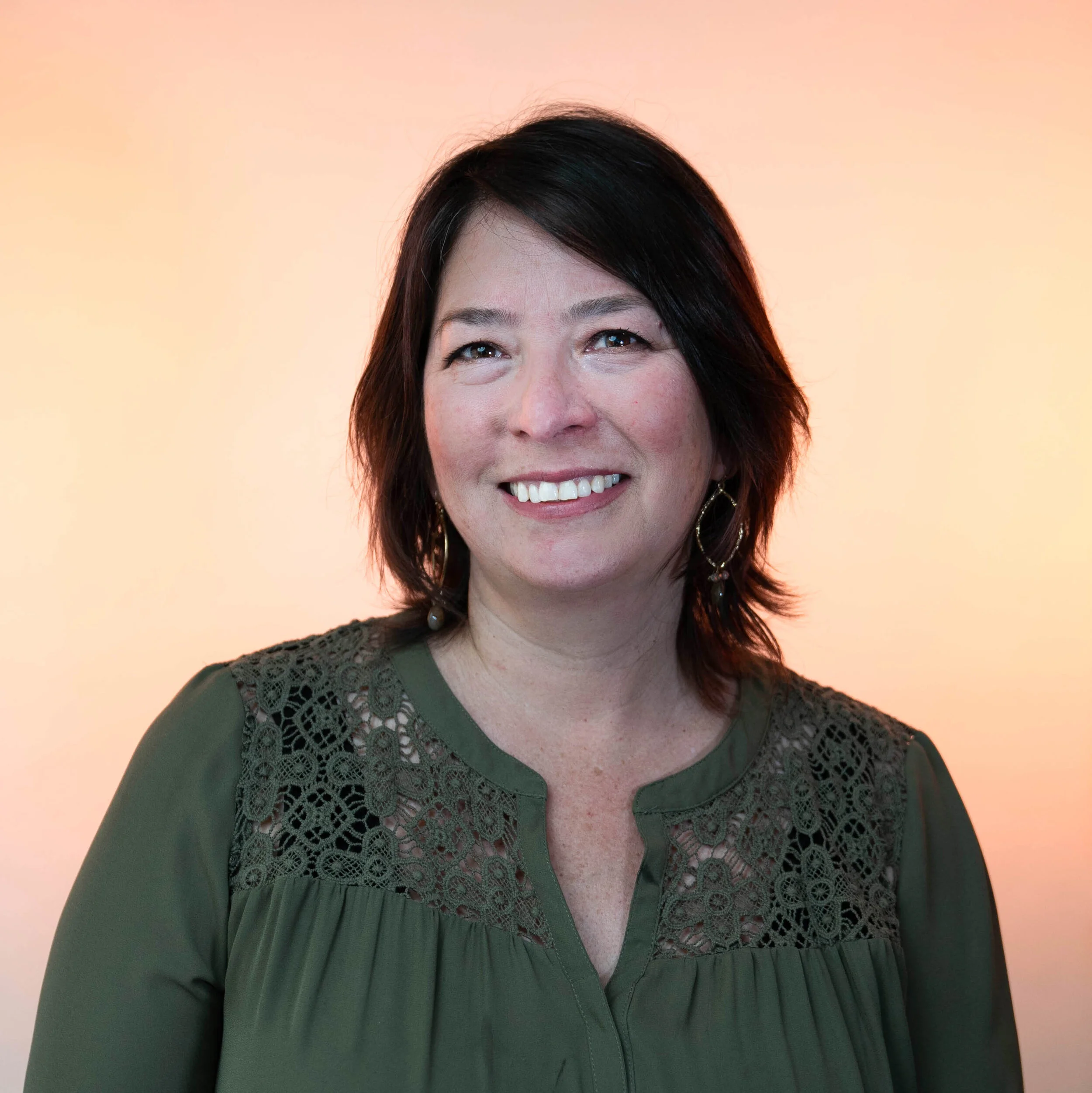 A Latina woman with shoulder length dark brown hair is smiling and wearing a dark green blouse with lace design and standing against a light orange background