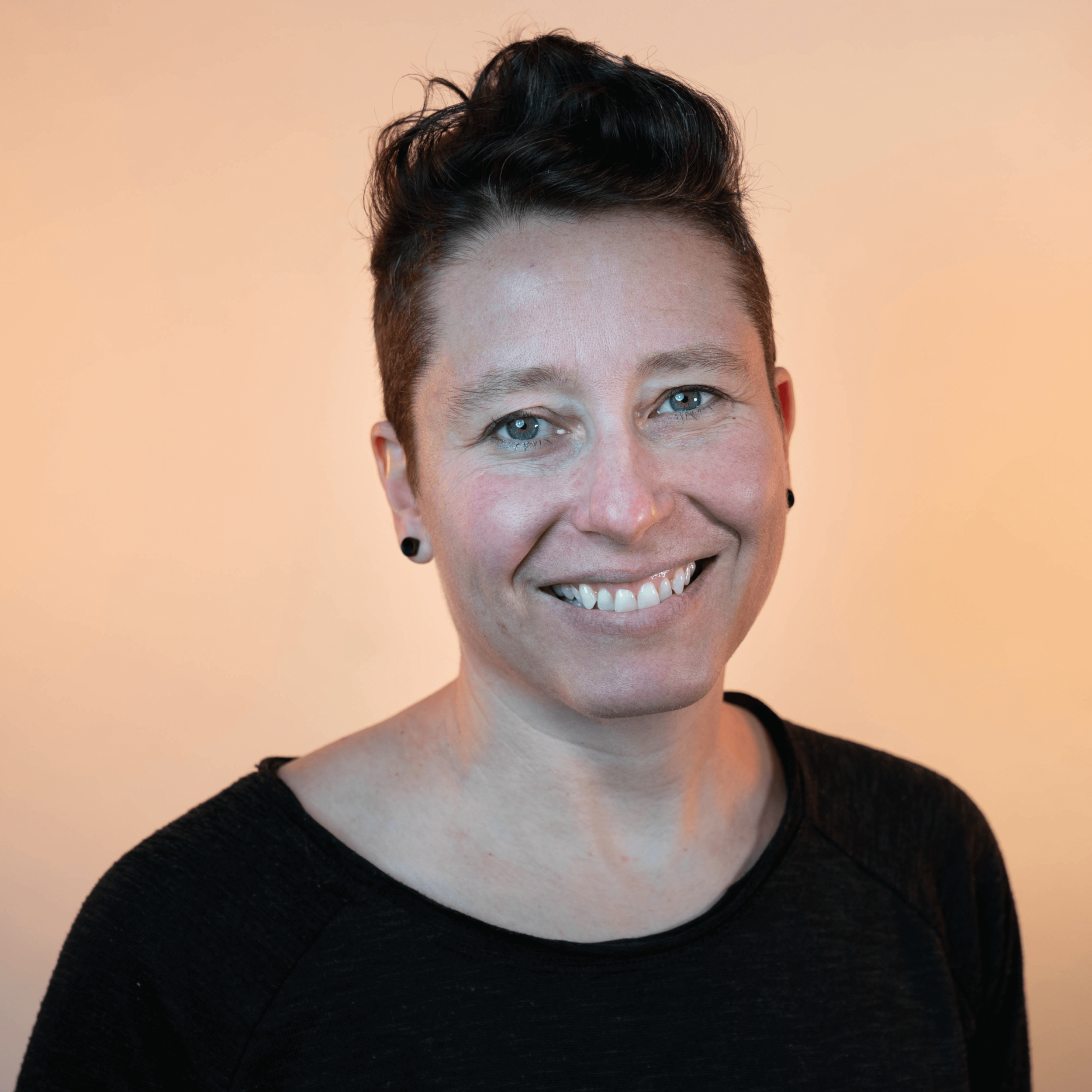 A white woman with short dark brown hair is smiling and wearing a black long sleeve shirt standing against a light orange background