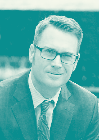Portrait of a man wearing glasses, a suit, and a tie, outdoors with a blurred background.