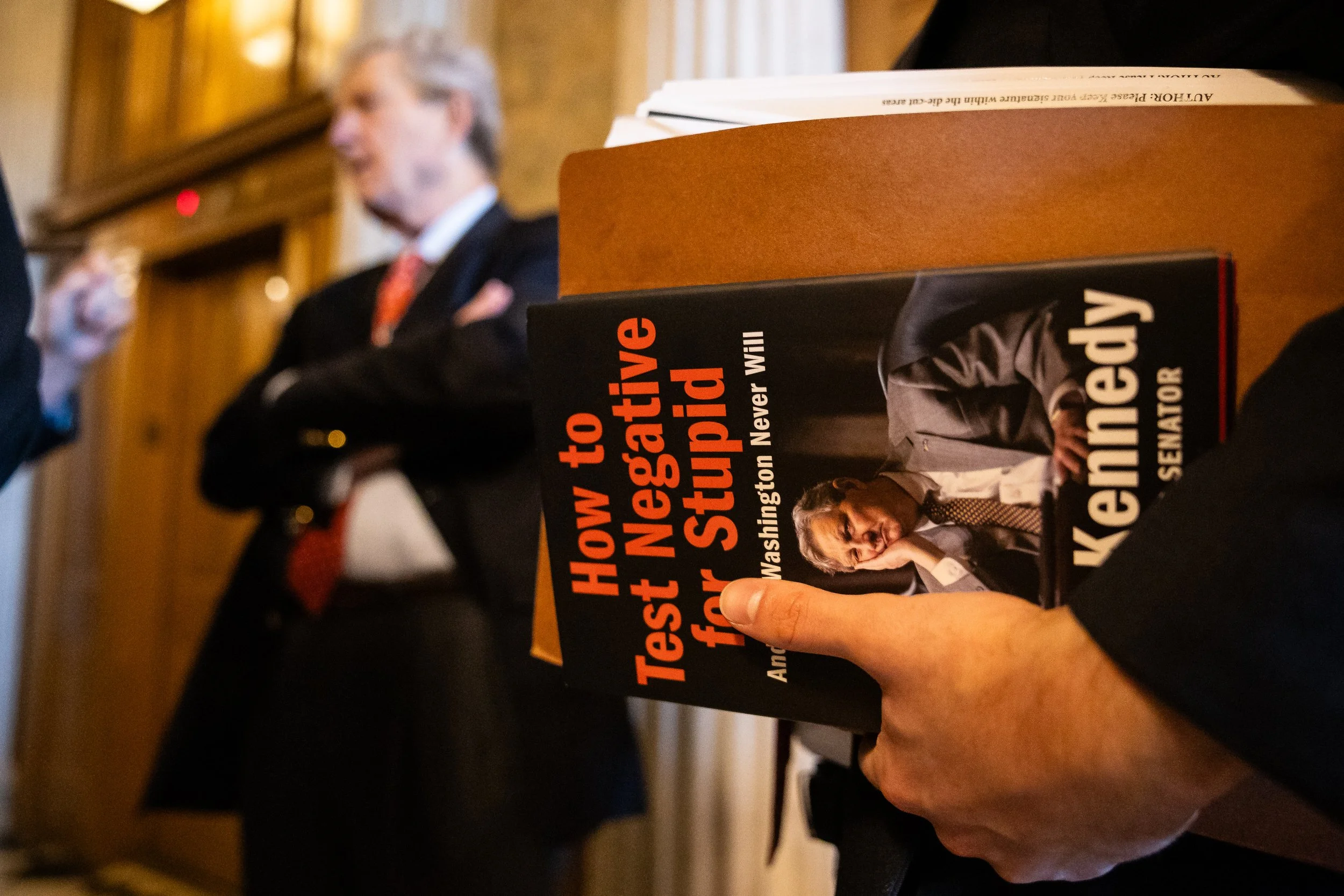  An aide holds a copy of "How to Test Negative for Stupid," a book by Sen. John Kennedy (R-La.), as Kennedy, at left, speaks with reporters after a vote at the U.S. Capitol on the 20th day of a government shutdown, Oct. 20, 2025.  