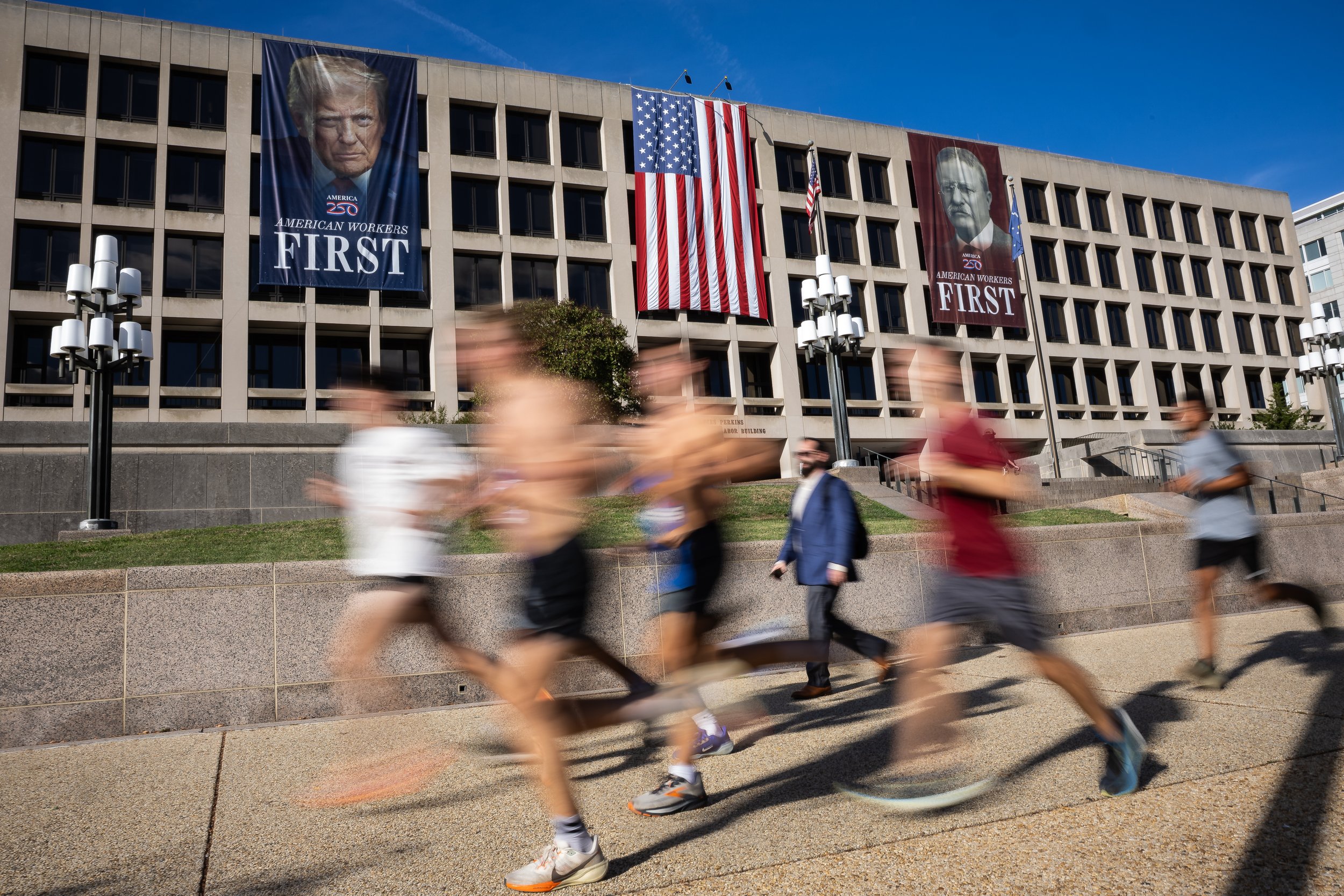  People run and walk past Department of Labor headquarters in Washington, D.C. on the second day of a government shutdown, Oct. 2, 2025. Images of Presidents Donald Trump and Theodore Roosevelt are seen on the building's exterior. 