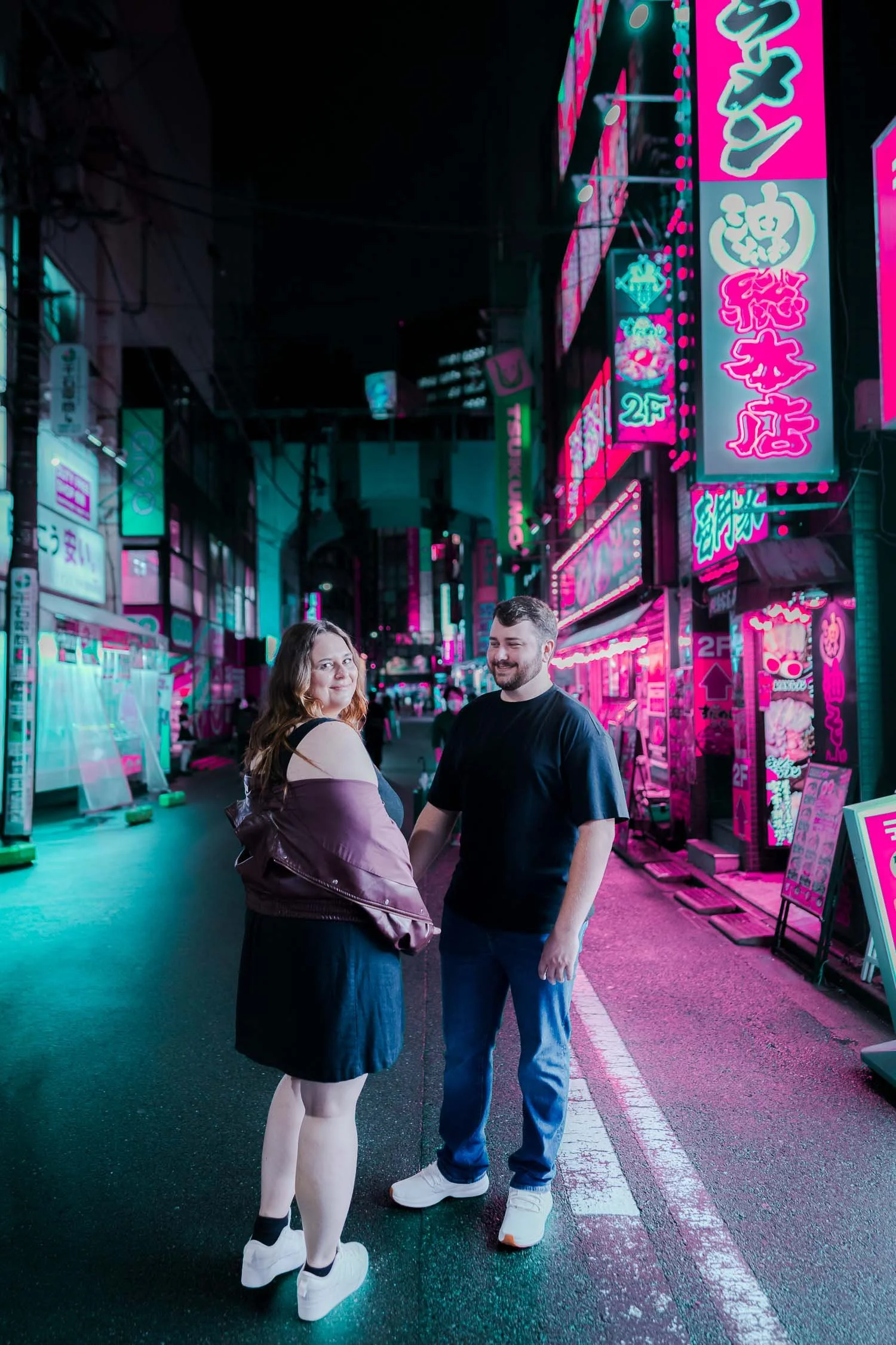 Couple pausing on a neon-lit Akihabara side street at night, glancing back at the camera during their Tokyo honeymoon photo session