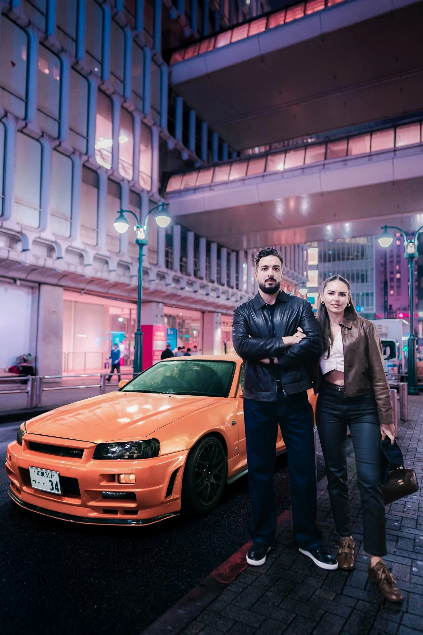 Couple standing in front of JDM, orange Nissan Skyline R34 during a Tokyo cyberpunk photoshoot in Shibuya
