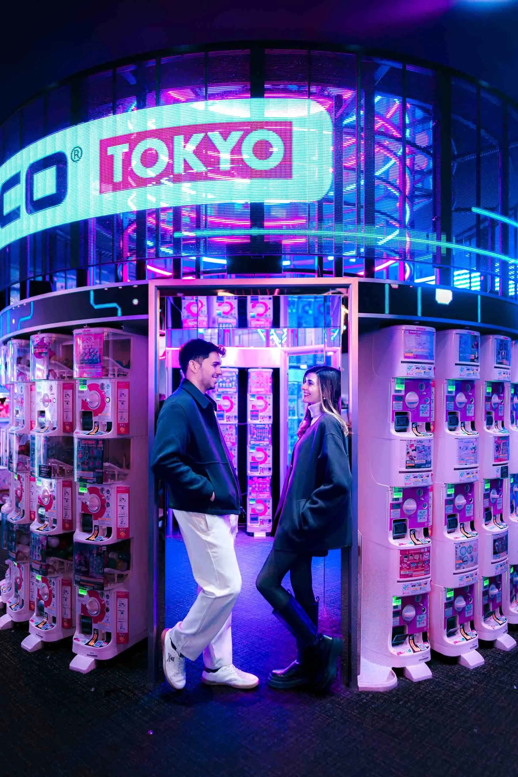 Couple standing and facing each other inside a colorful Shinjuku capsule toy arcade, surrounded by rows of gachapon machines and vibrant cyberpunk neon lighting.