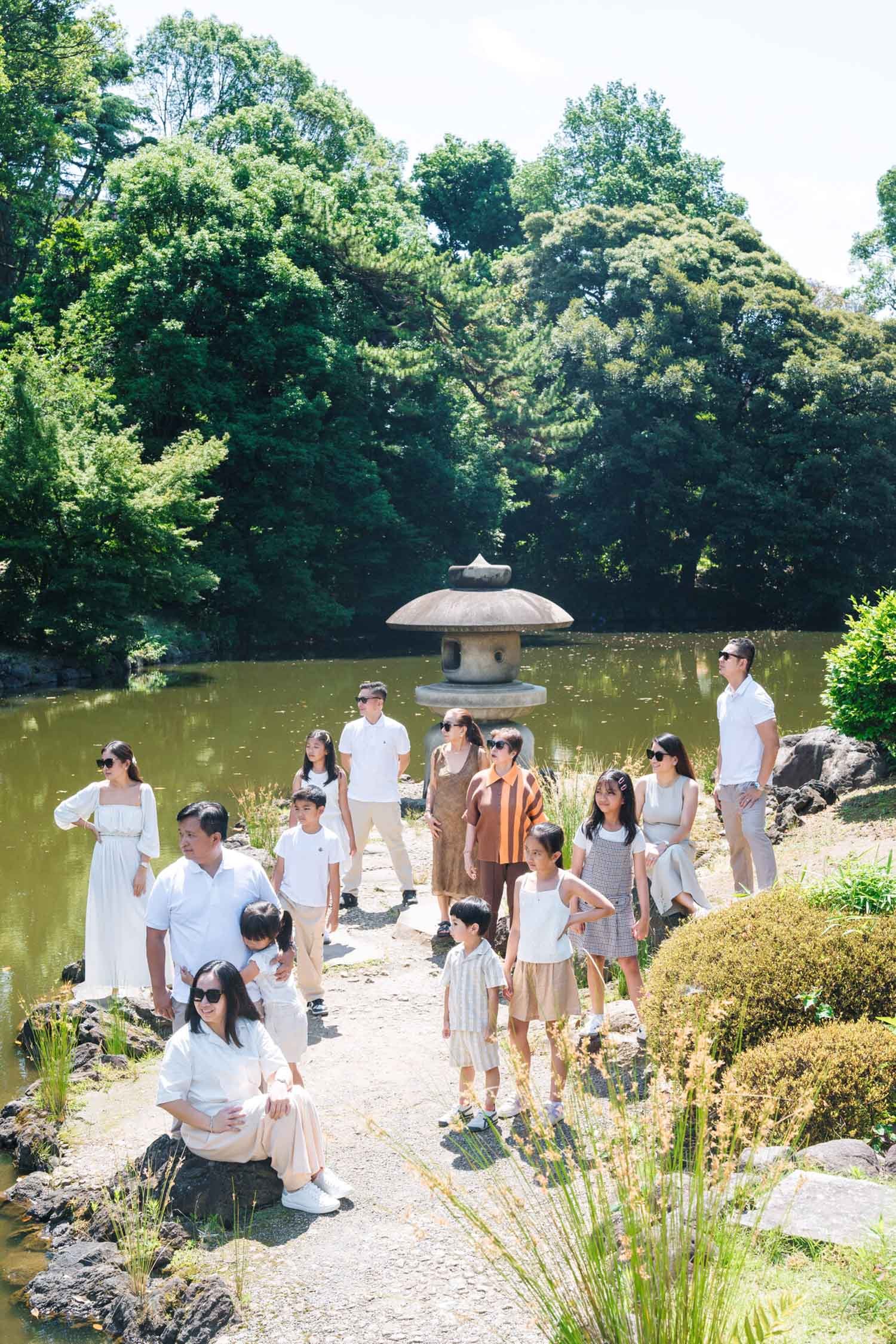 Large extended family in coordinated neutral outfits beside a pond and stone lantern in a Tokyo garden