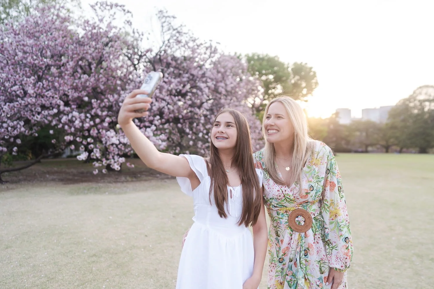 Mother and daughter taking their selfie in front of cherry blossom trees with golden sunset in the background.