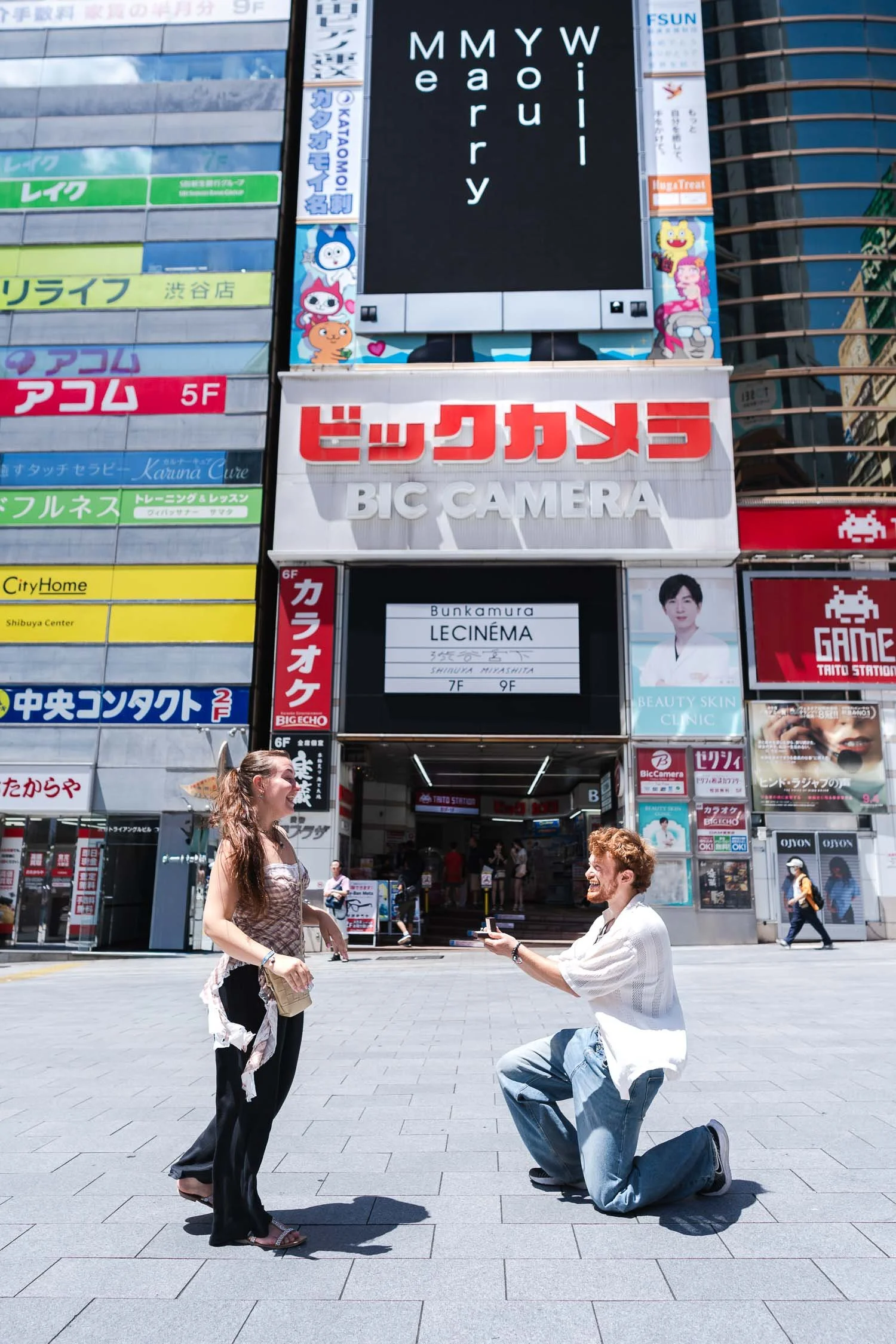 Man kneeling to propose in front of Shibuya digital billboard displaying 'Will You Marry Me' message above Bic Camera store