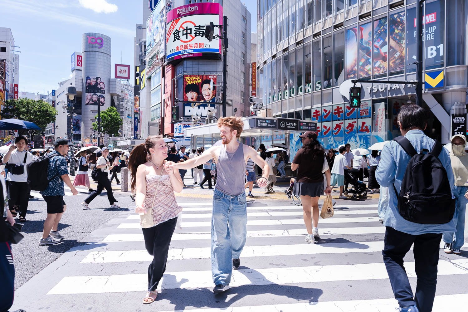 Engaged couple walking hand in hand across Shibuya Crossing with Tokyo skyline and billboards in background