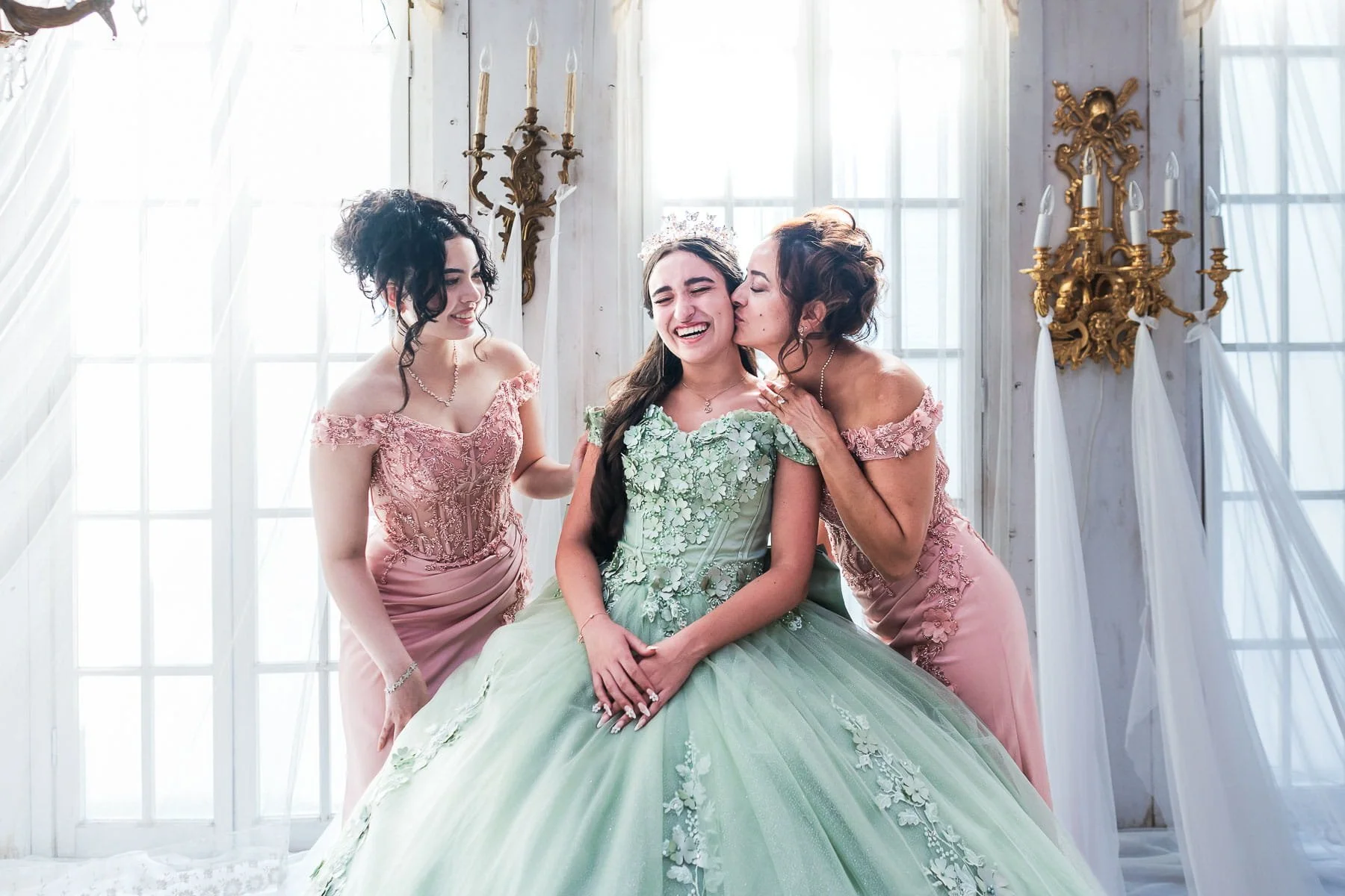 Quinceañera in a mint green ball gown laughing as her mother and sister in pink dresses embrace her, indoor studio photoshoot in Tokyo