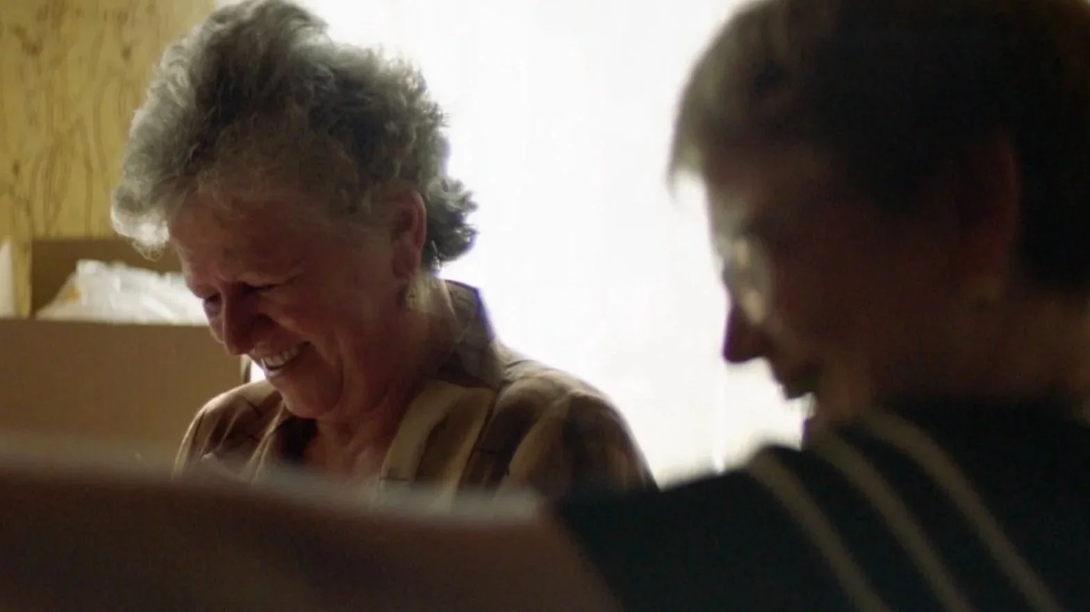 Some elderly Romanian women are laughing while kniting in front of a w9ndow so they are partially silhouetted