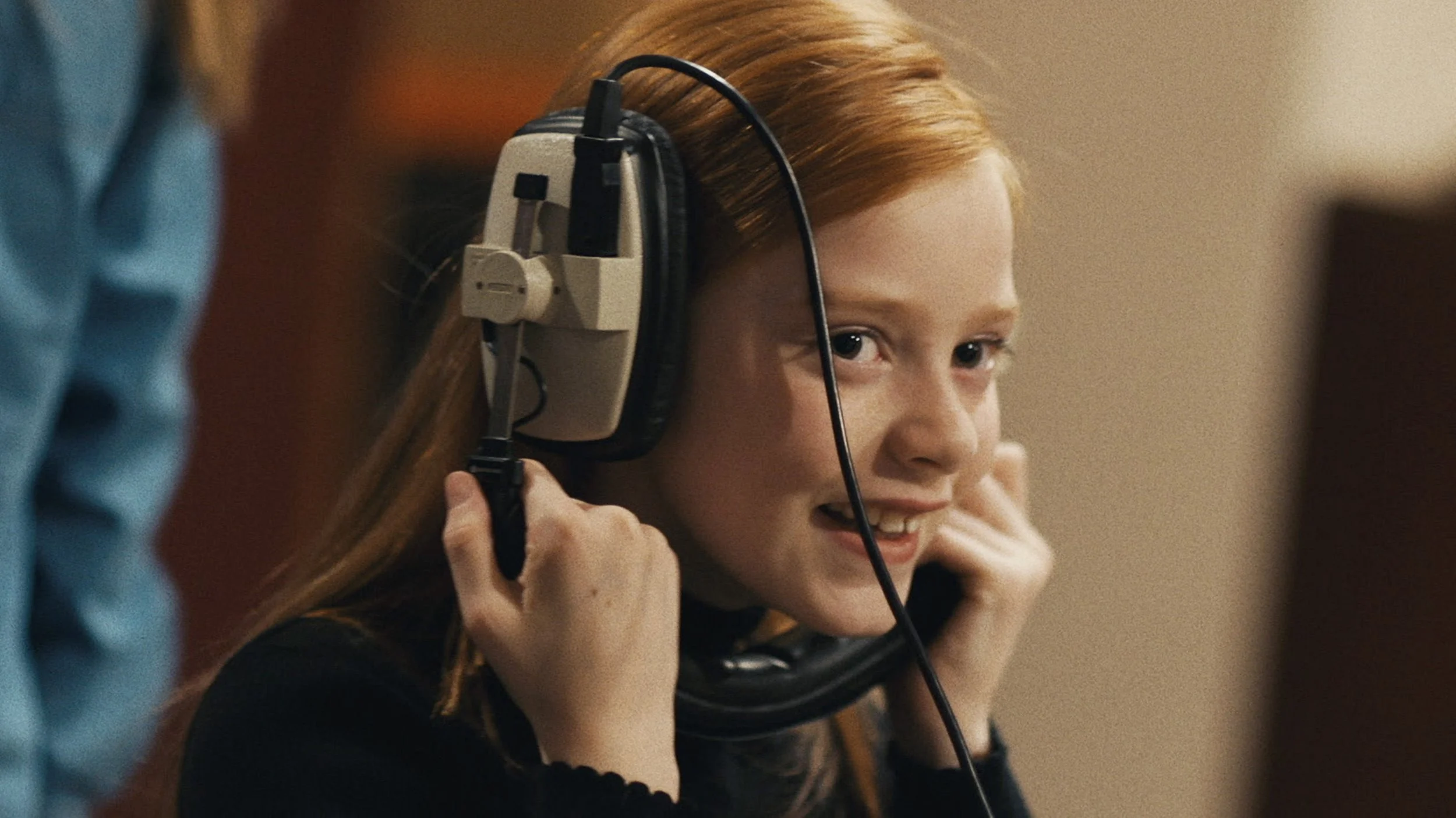 A young girl with ginger hair is smiling while holding sound recording studio headphones over her ears upside down
