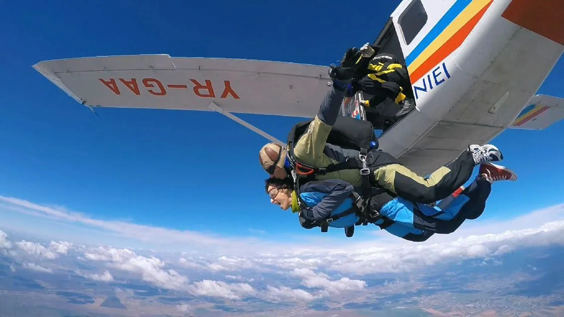 We see a young Romanian woman jump out of a small plane in a tandem skydive. She is screaming, and the view looking down into the clouds is epic