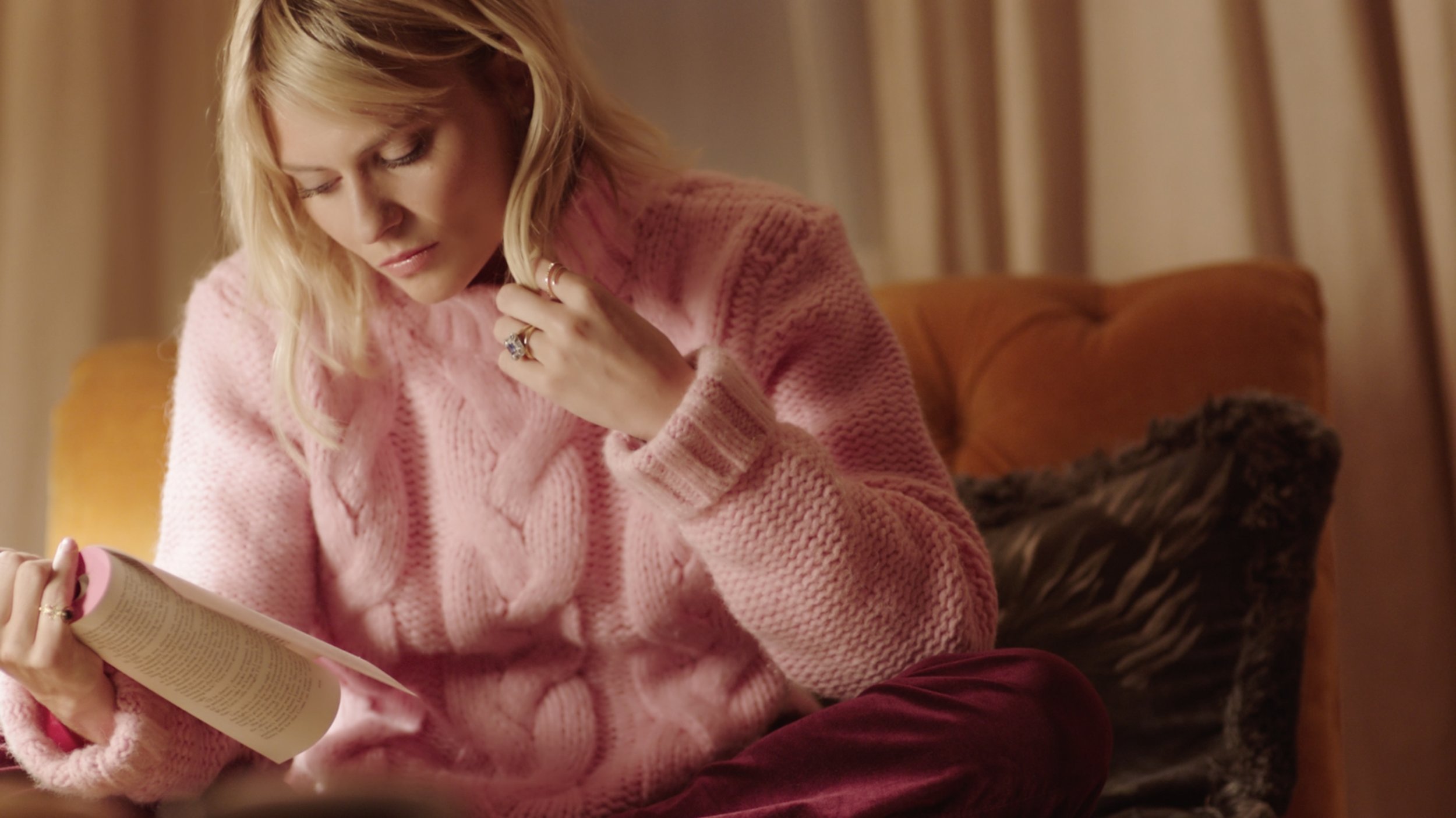 A woman, Pandora Sykes, is sitting in a comfortable armchir reading a book. She is playing with her hair with her spare hand and weraing a bright pink mohair jumper