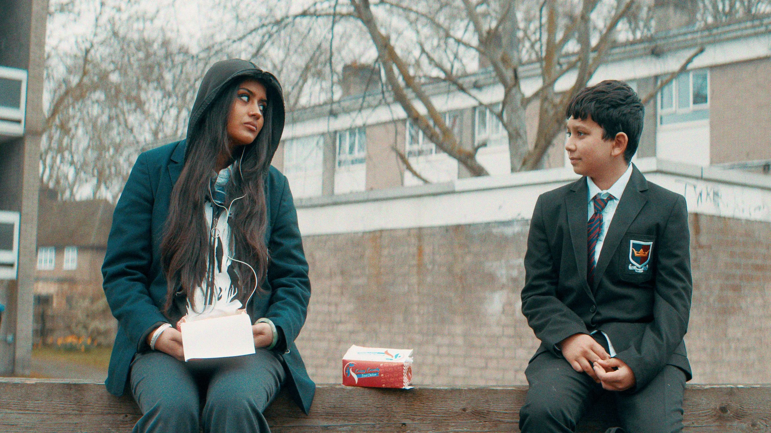 Two schoolchildren - one male, one female, are sitting on a low wall in a London suburban street. They are wearing school uniform and have boxes of chicken and chips from a takeaway. They don't look like they are very friendly