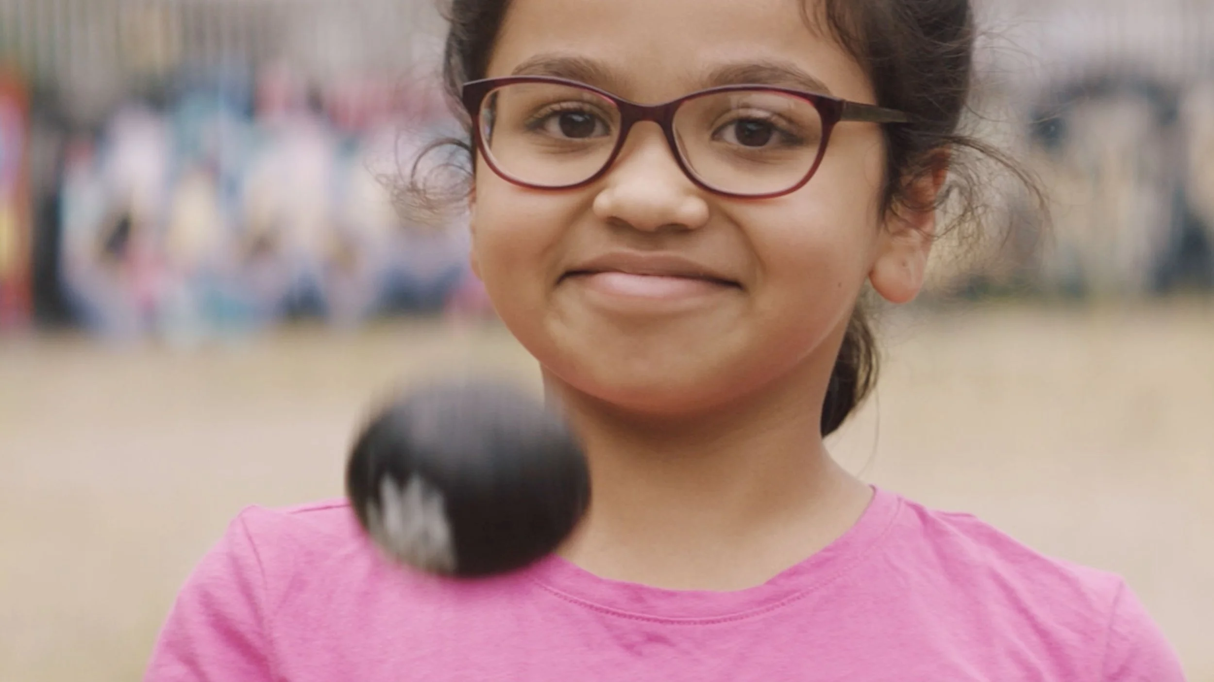 A young South Asian girl with glasses is smiling to camera and throwing a cricket ball up