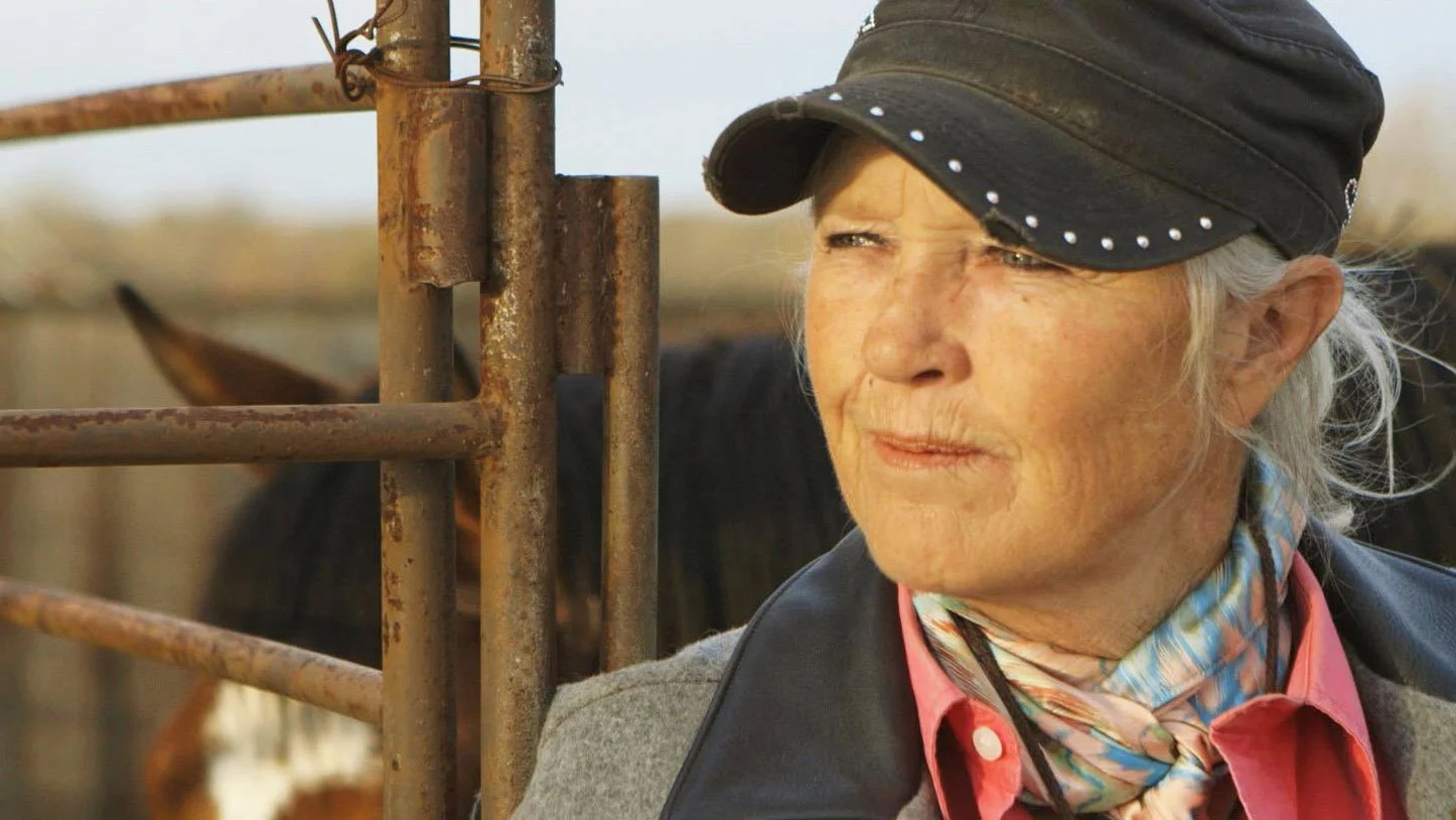 An elderly woman dressed in western rodeo style clothing is smiling as she looks out to the distance. In the back ground we see a horse in a pen