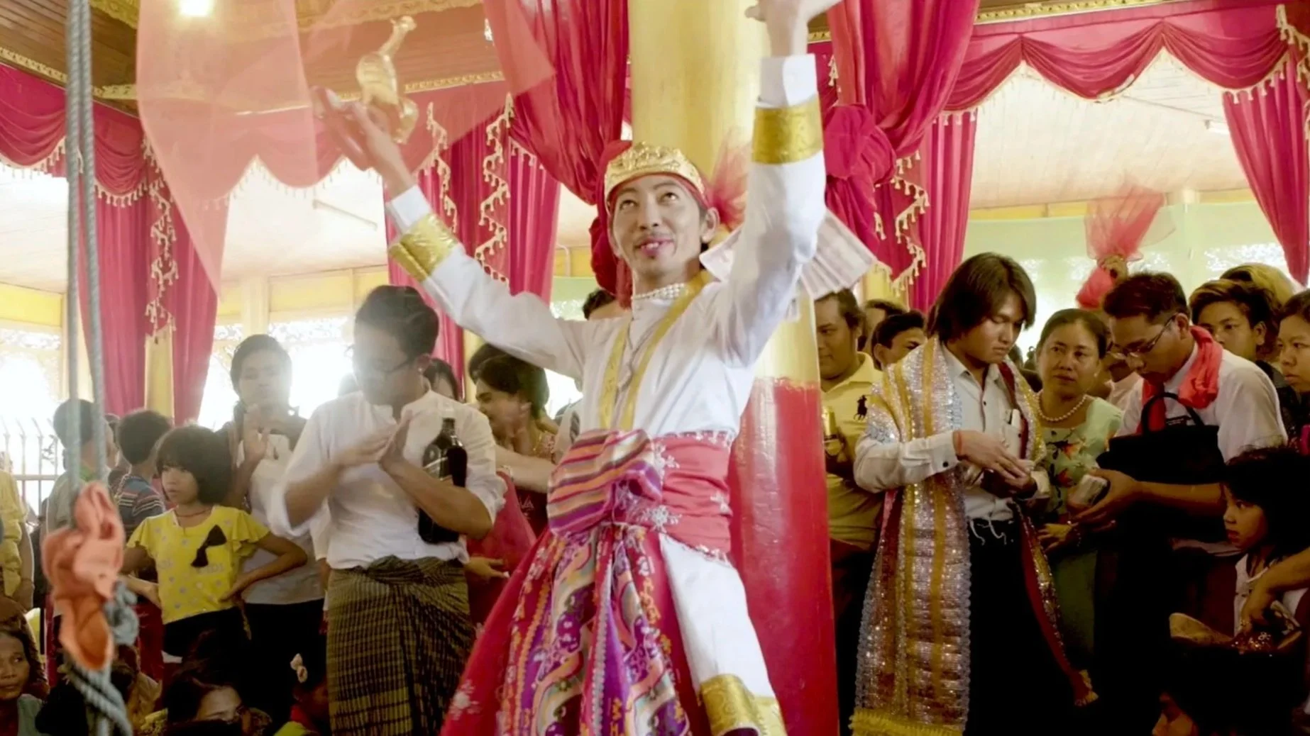 An image from a religious ferstival in Myanmar. A man dressed in bright robes and festive fabric and wearing a gold crown is dancing with his hands in the air. Around him a crowd of families are throwing moneyy