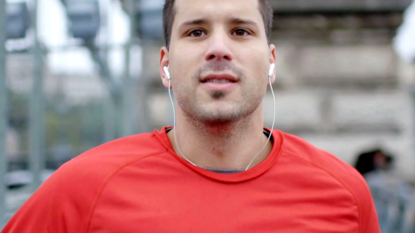 A young man in a red top is running toward camera listing to his airpods. We can see he is crossing an industrial style bridge