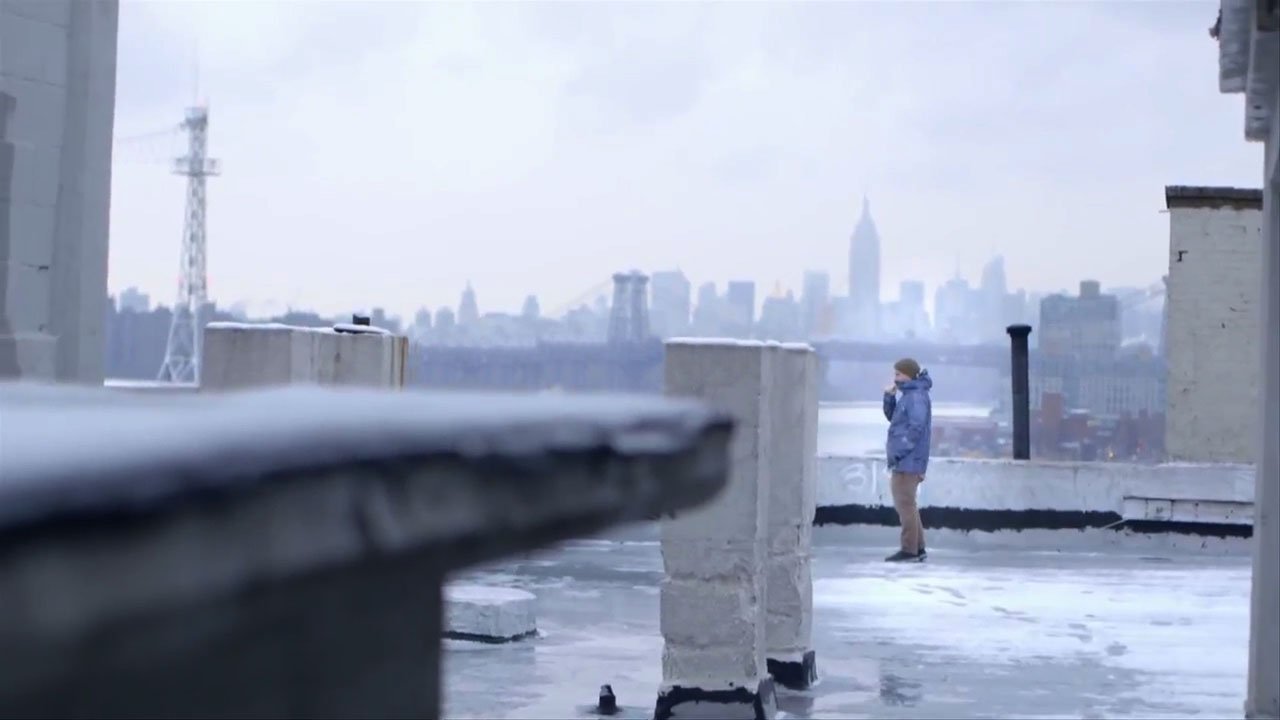 An artist - Zach Lieberman - is standing on the roof of an industrial building in Brooklyn with the Manhattan skyline in the background. There is snow on the ground and we see his breath