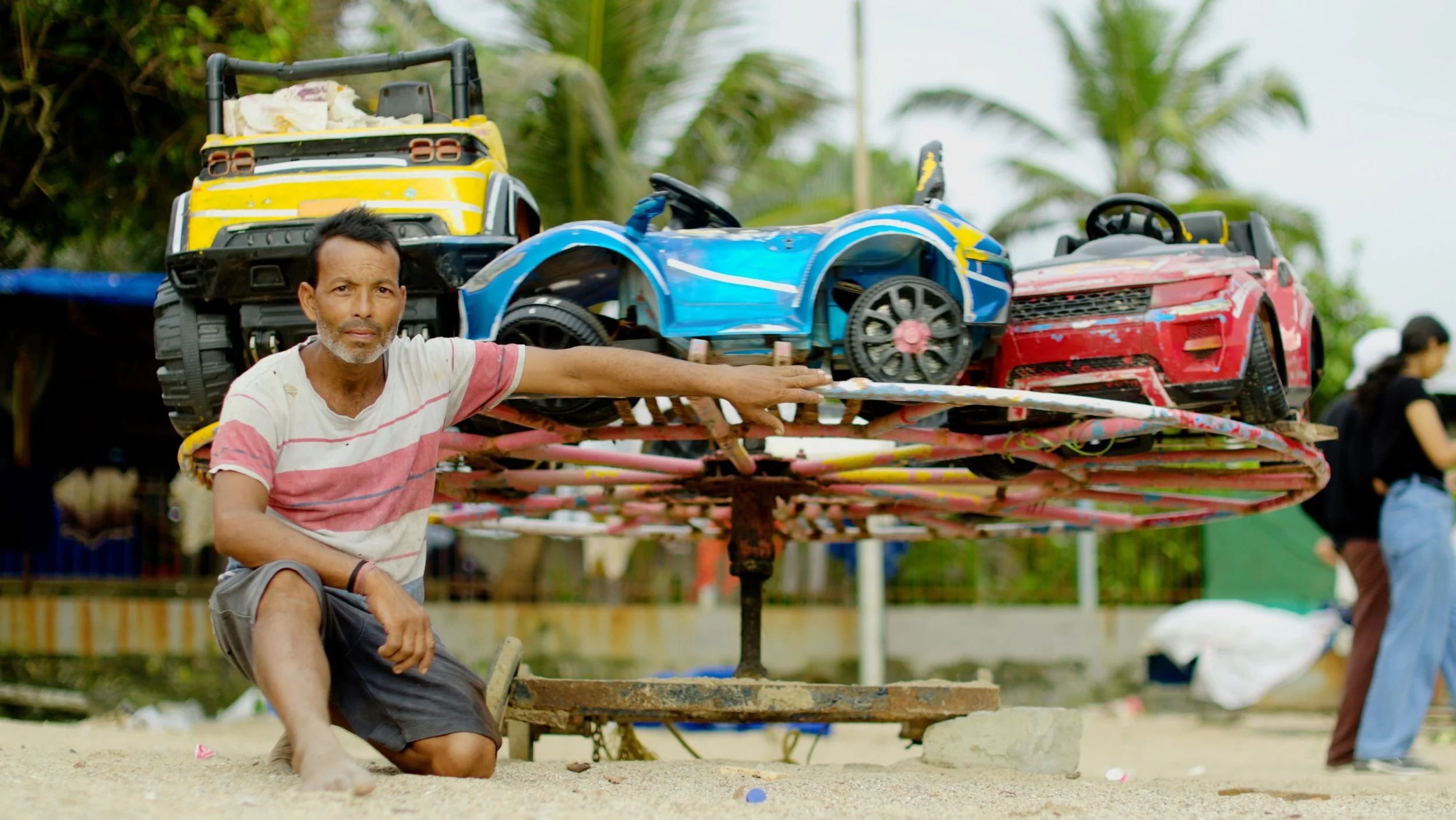 A middle aged Indian man poses next to a brightly coloured childrens roundabout on a beach. There are palm trees in the background and he is wearing shorts and a tee shirt