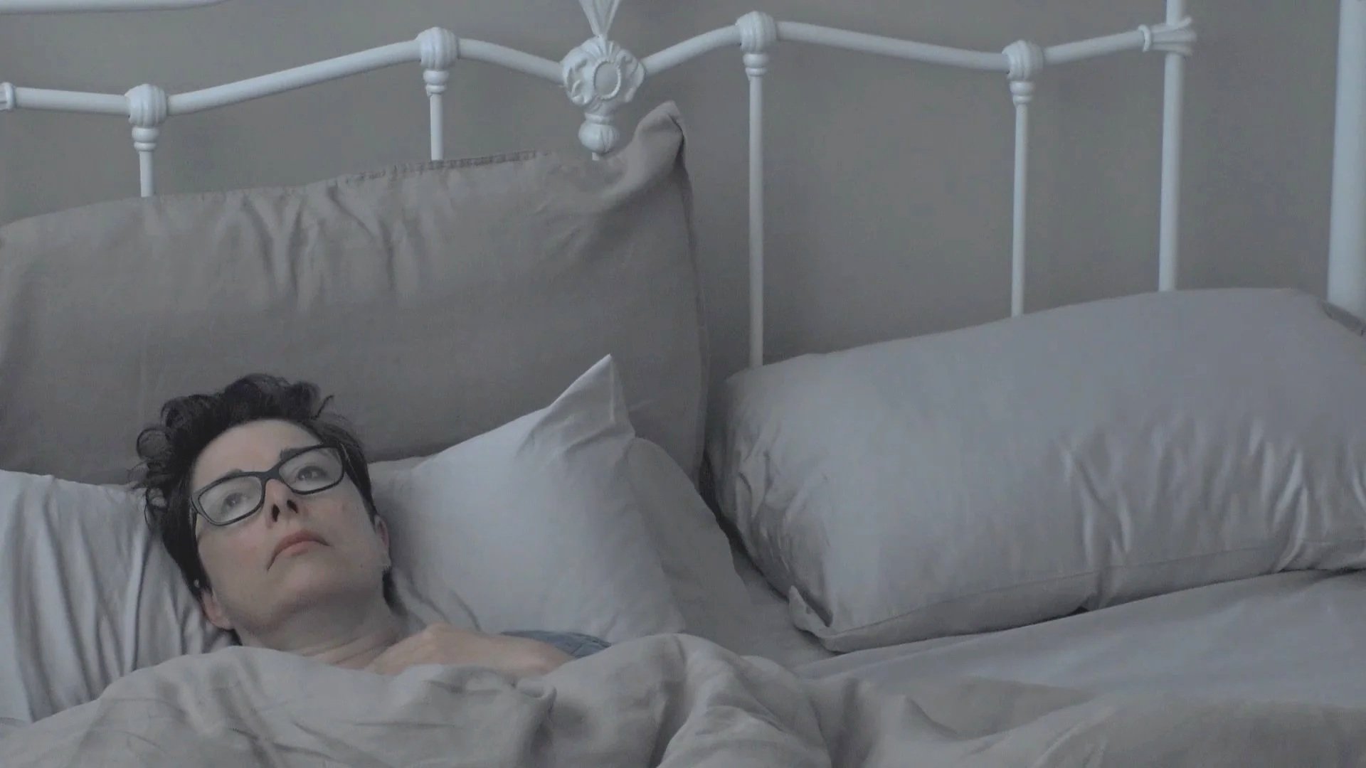 A woman with glasses - Sue Perkins - in lying in a double bed looking reflective and lonely