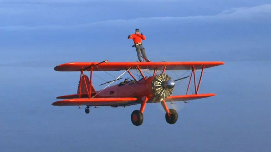Against a blue sky a red biplane is cruising through the aie with a man standing on the top doing a 'wing walk'. he is wearing a red top and sunglasses