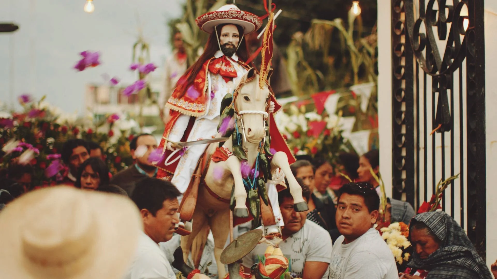 A wooden carved and brightly painted statue of a catholic saint riding a horse, is being carried through a crowd by some Mexican men as part of a religious festival