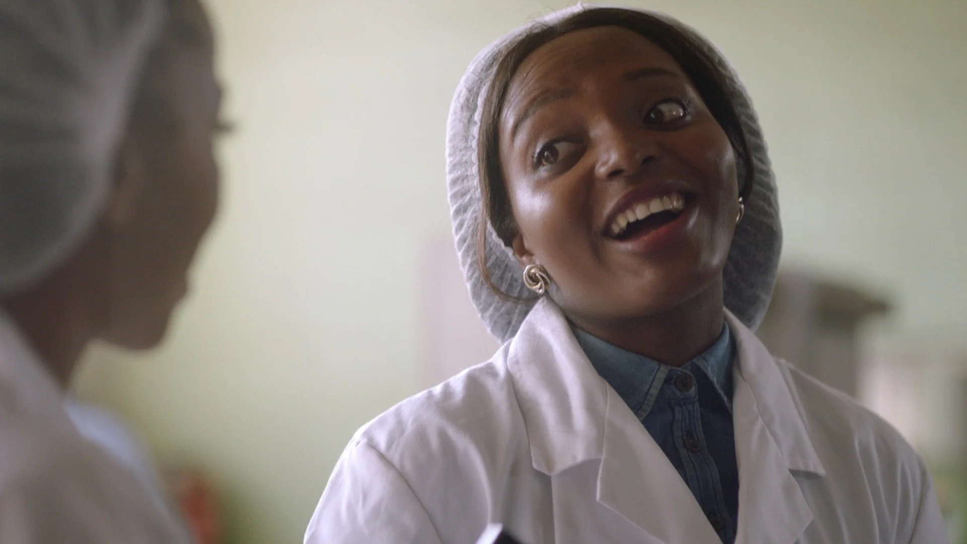 A factory line worker in white overalls and a hair net is laughing chatting to another woman. Both are black