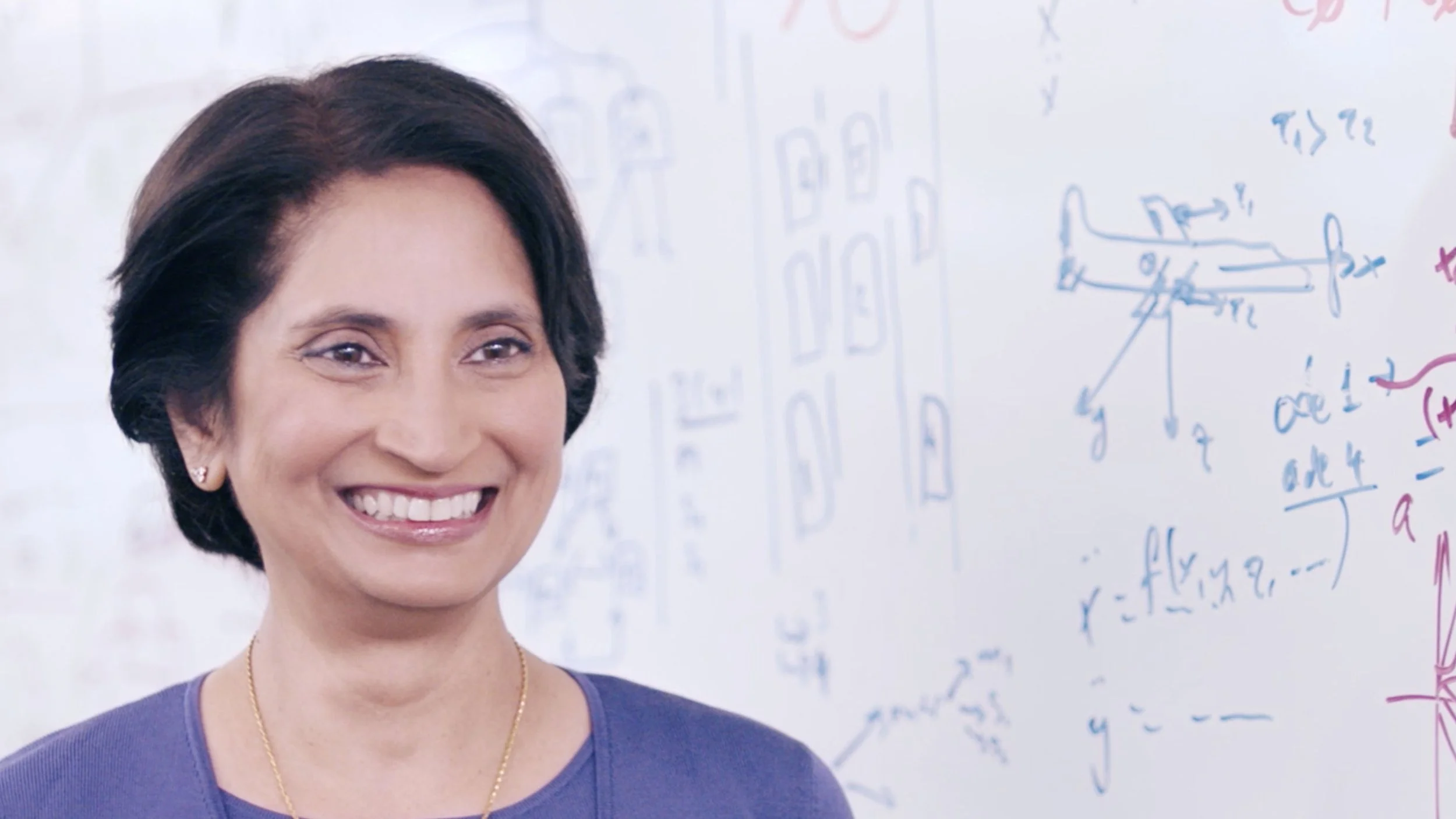 A middle aged Indian woman - CEO Padmashree Warrior - is standing infront of a white bopard covered with formulars and smiling
