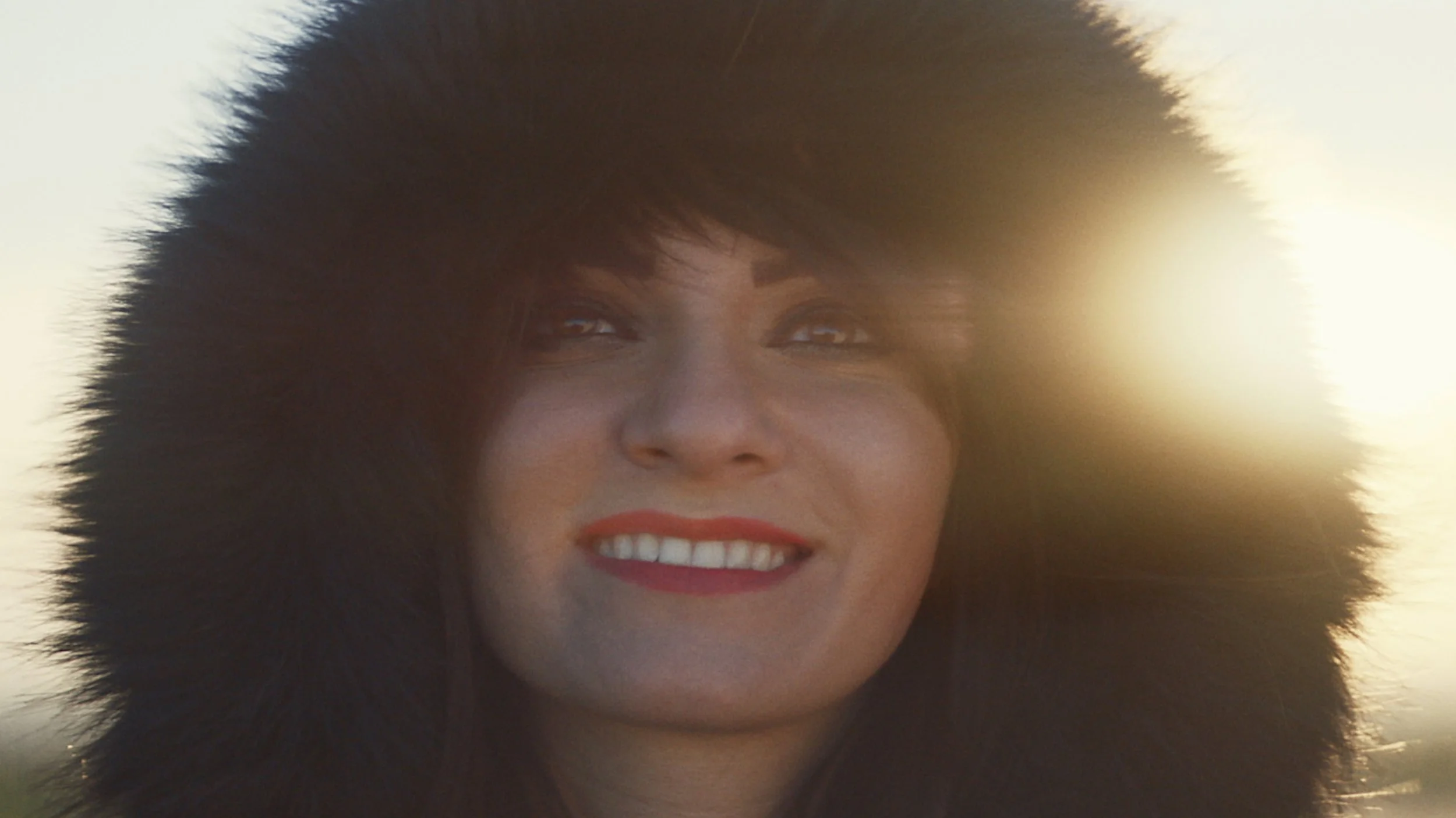 A young woman is wearing a very warm looking furry parker with the hood pulled up. She is smiling and the sun behind her is flaring into the lens