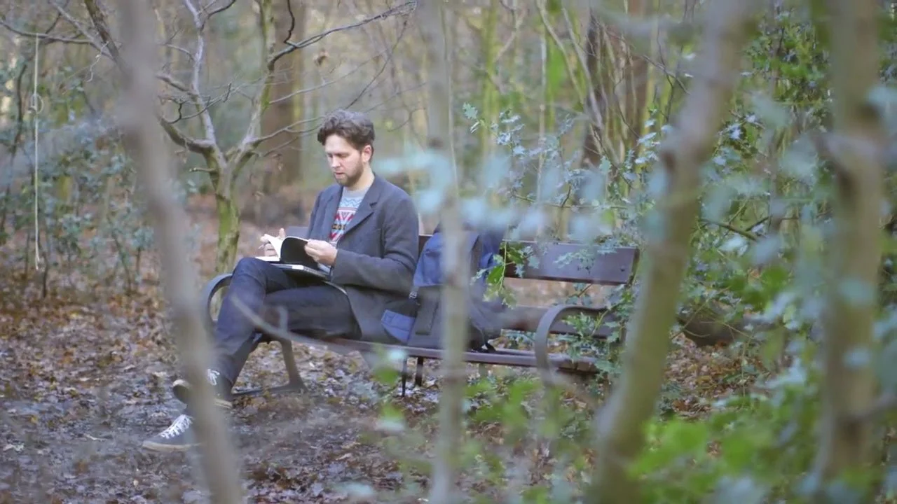 A white male in his 30s is sitting on a bench in the woods with his laptoip on his lap and he is looking through his notebook