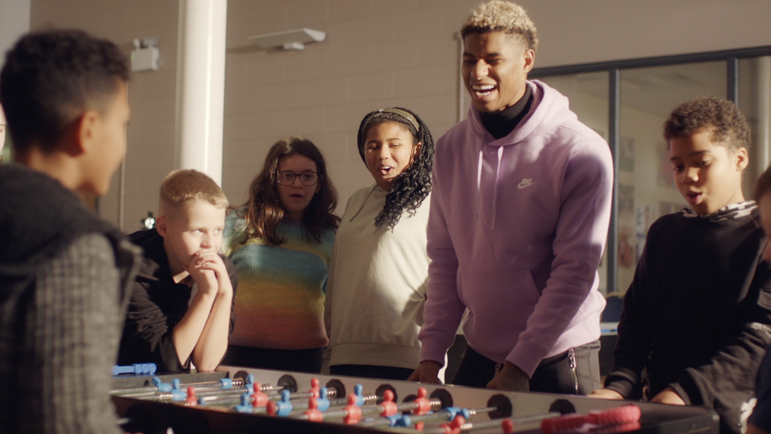 A group of 5 children are standing round a table football game enjoying the game, while Premiership footballer Marcus Rashford celebrates scoring a goal