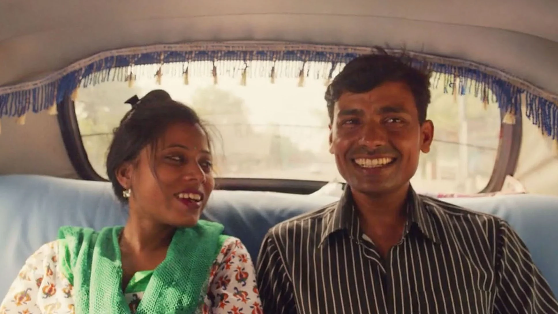 An Indian man and woman are smiling and joking with each other while riding in the back of a classic Delhi black taxi with decorative fringing on the ceiling