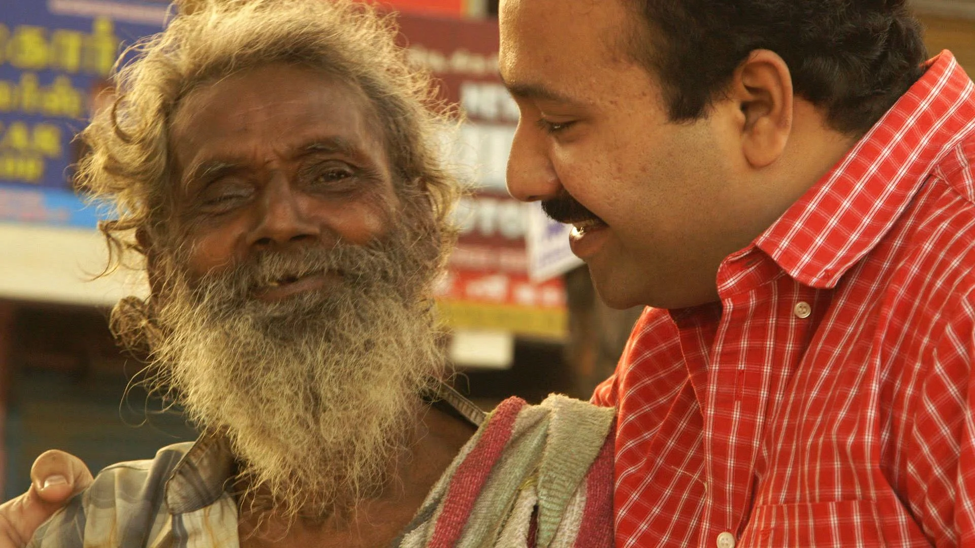 A kind looking Indian man is smiling while he puts his arm around an older Indian man's shoulder. The older man is unkempt, with hair overgrown as if he is homeless