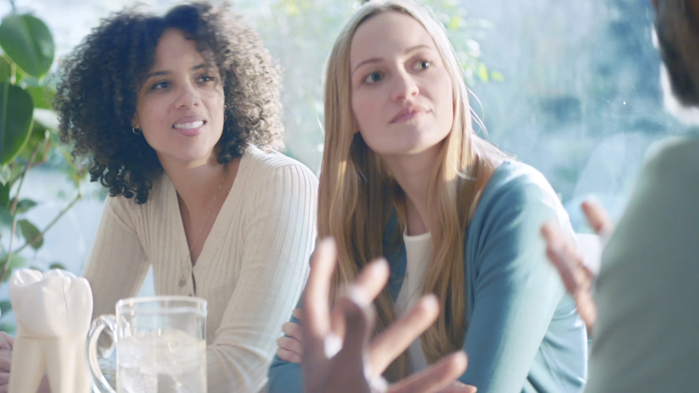 Two women are showing interest in what another person off camera is telling them about tooth sensitivity. Sun is streaming through the window behind them