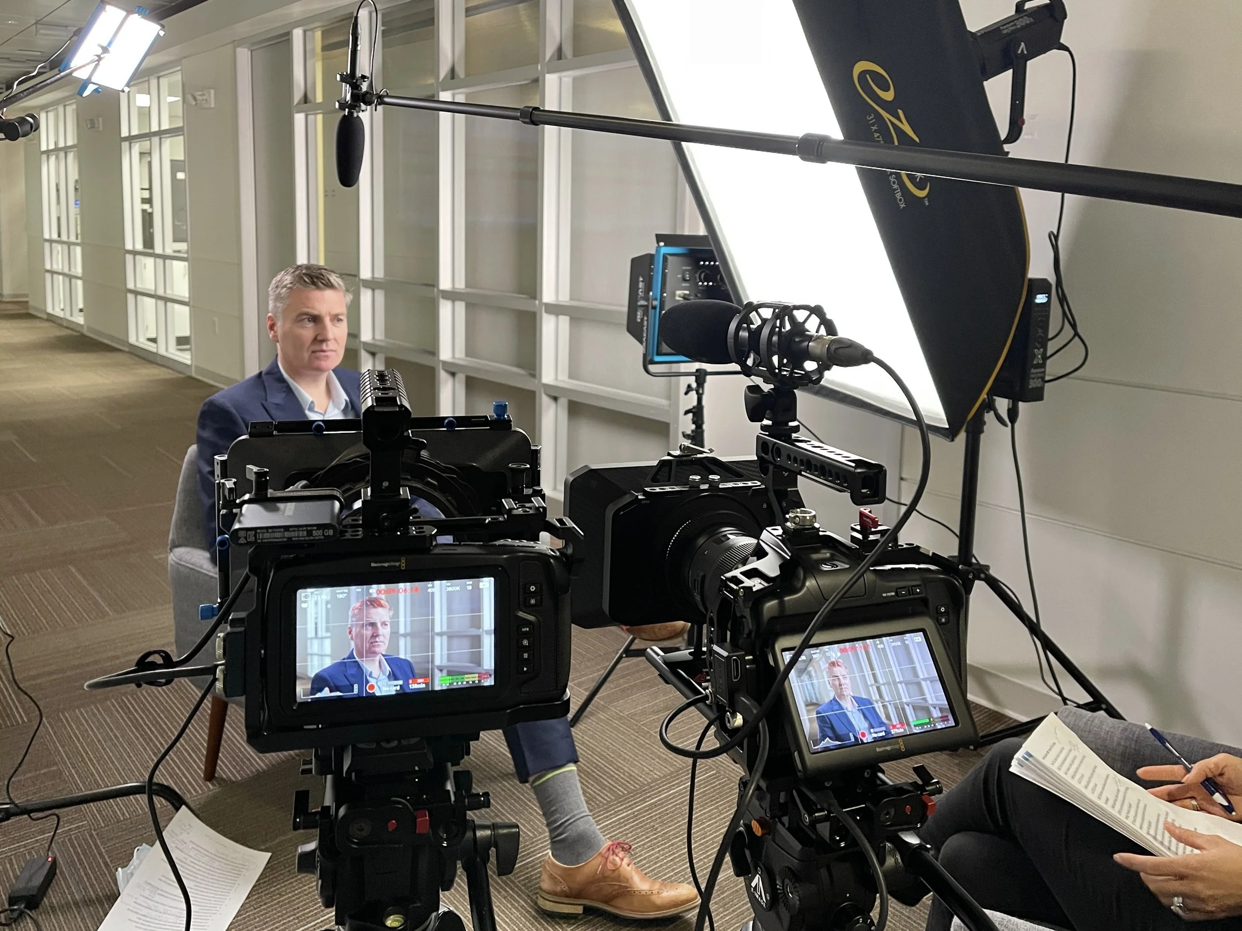 A man in a suit being filmed in an interview setting with cameras, lights, and crew, inside a modern office or conference area.