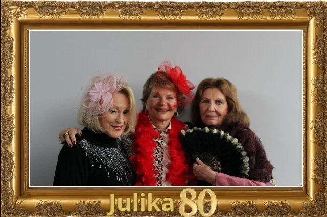Tres mujeres mayores vestidas con ropa elegante y decoradas con accesorios coloridos, posando juntas en un marco dorado con la inscripción 'Julika 80'.