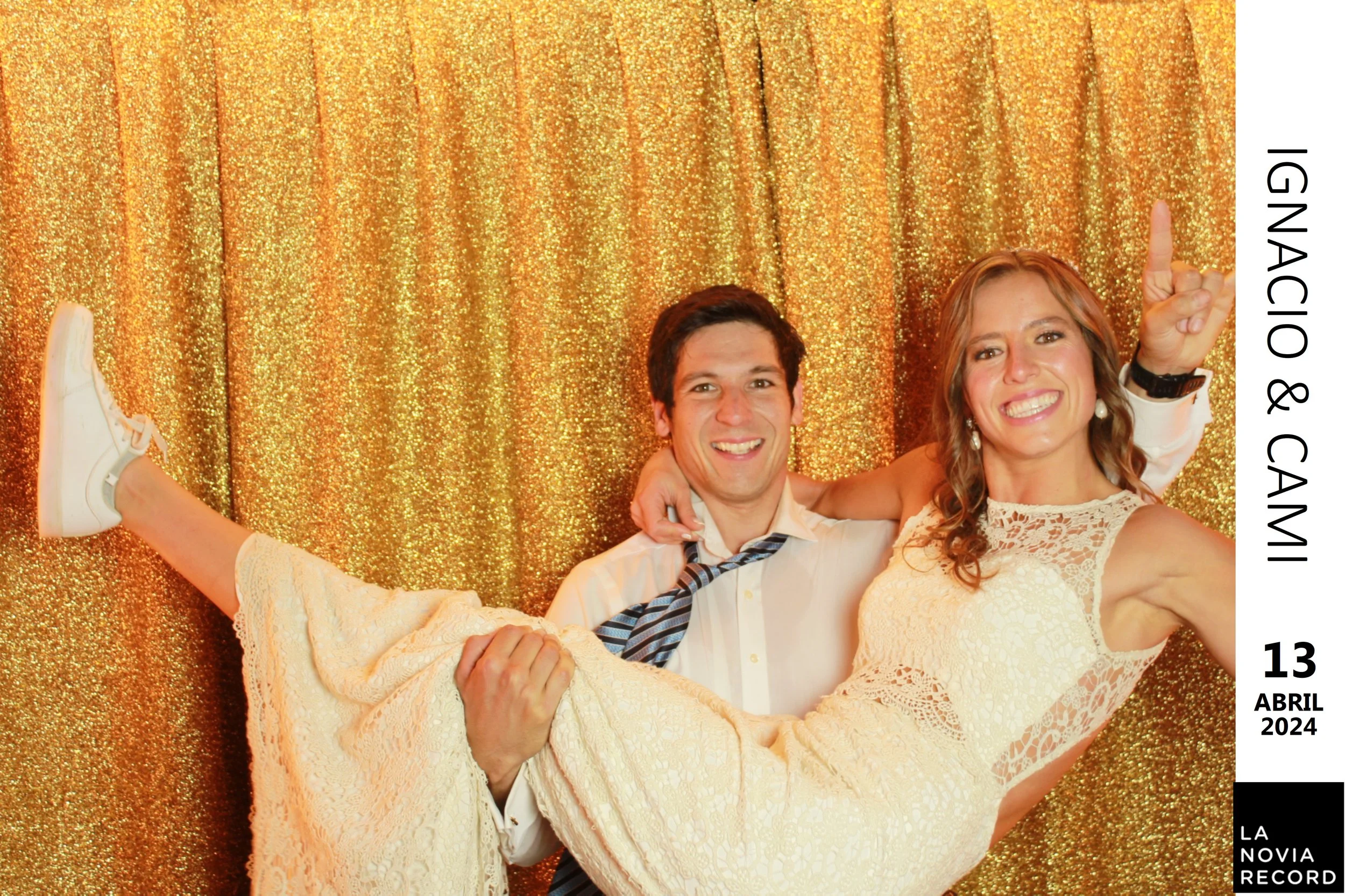 Una pareja sonriente en un fotomatón con fondo dorado, la mujer lleva un vestido blanco y el hombre una camisa blanca con corbata a rayas, él la sostiene en brazos mientras ella abraza con una mano levantada.