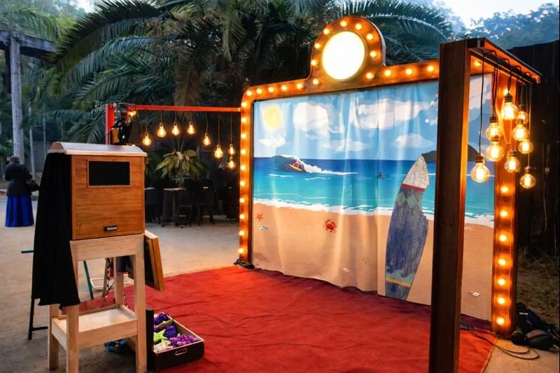Escenario decorado con tema de playa, con cortina de tela con imagen de la playa, luces colgantes, y fondo con palmeras y personas en la playa.