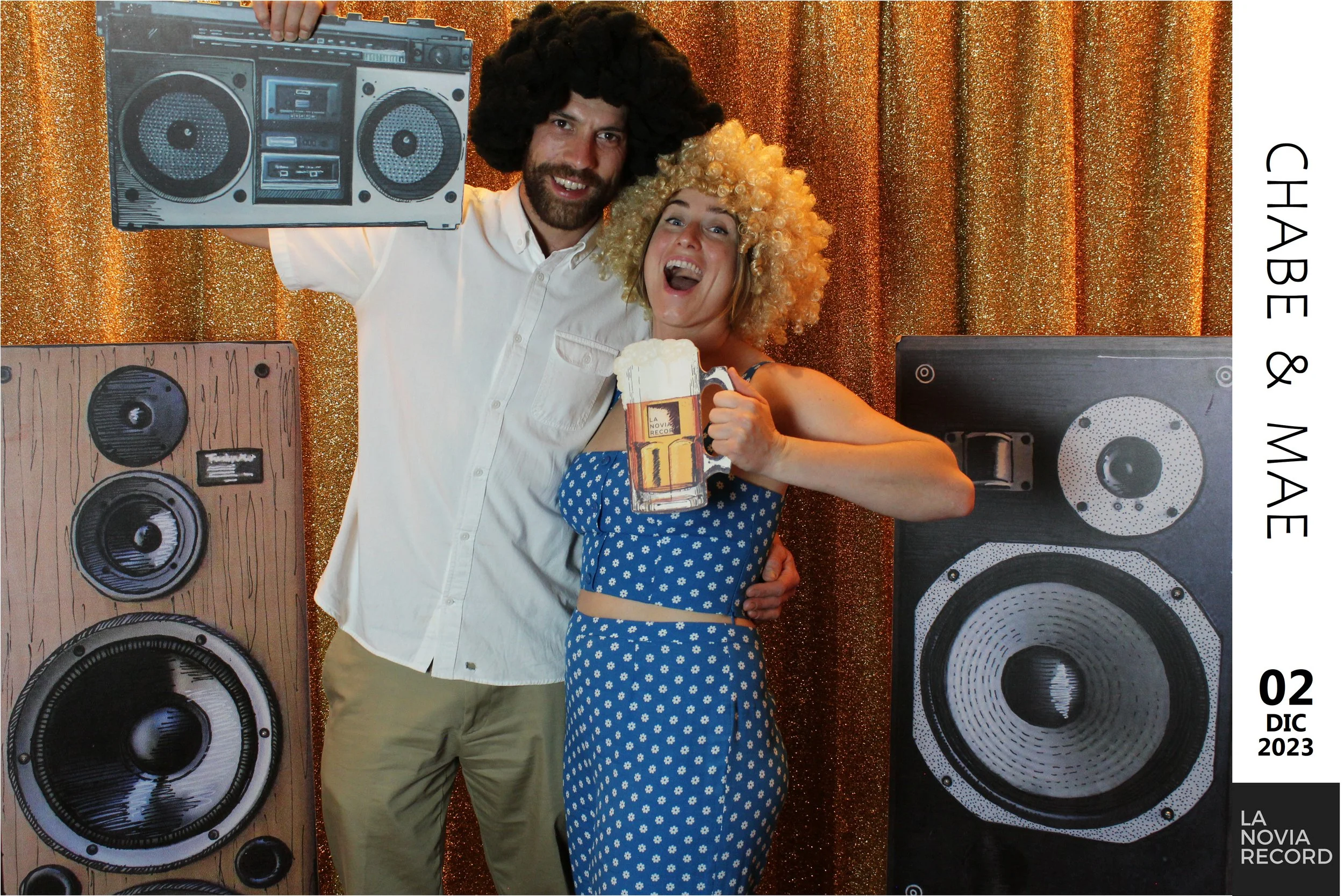 Una pareja feliz en una fiesta con decoraciones doradas, vestidos y accesorios retro; tienen grandes altavoces y un fondo de cortina dorada. Ella sostiene un cartel de cerveza y él sostiene un cartel de un equipo de música.
