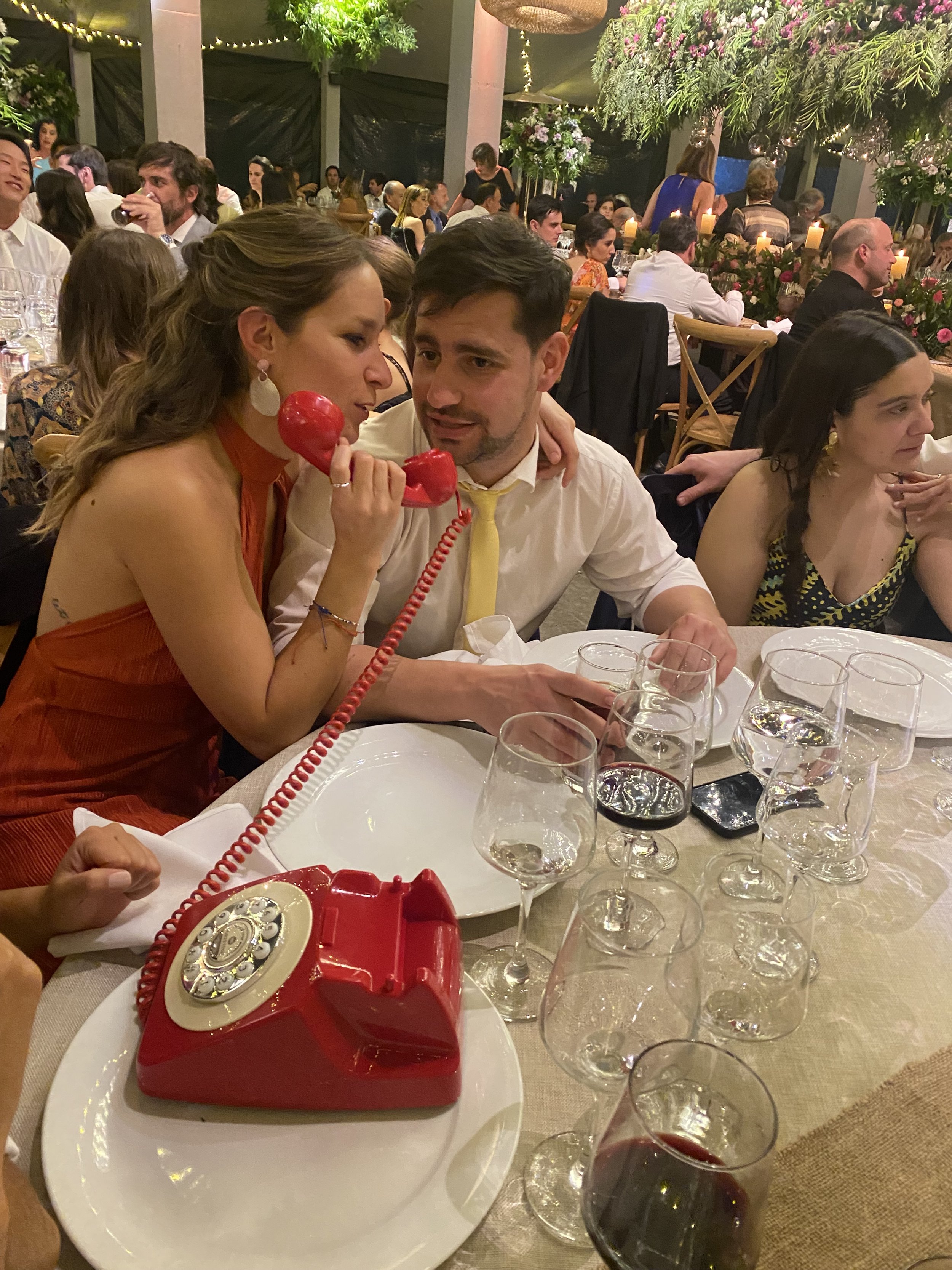 Personas en una fiesta sentadas en una mesa, una mujer con vestido naranja y aretes grandes hablando por teléfono rojo, y un hombre con camisa blanca y corbata amarilla, en un ambiente con muchas personas, decoraciones florales y velas.