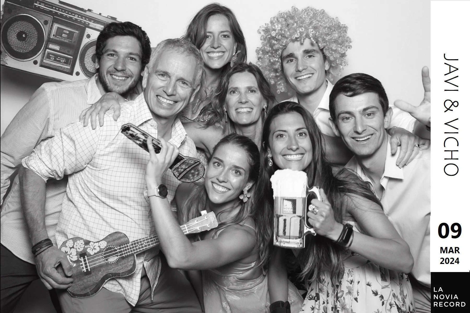 Un grupo de personas en una fiesta, sonriendo y posando con bebidas y una guitarra, en un fondo de equipo de sonido, en blanco y negro, con fecha y evento en la esquina derecha.