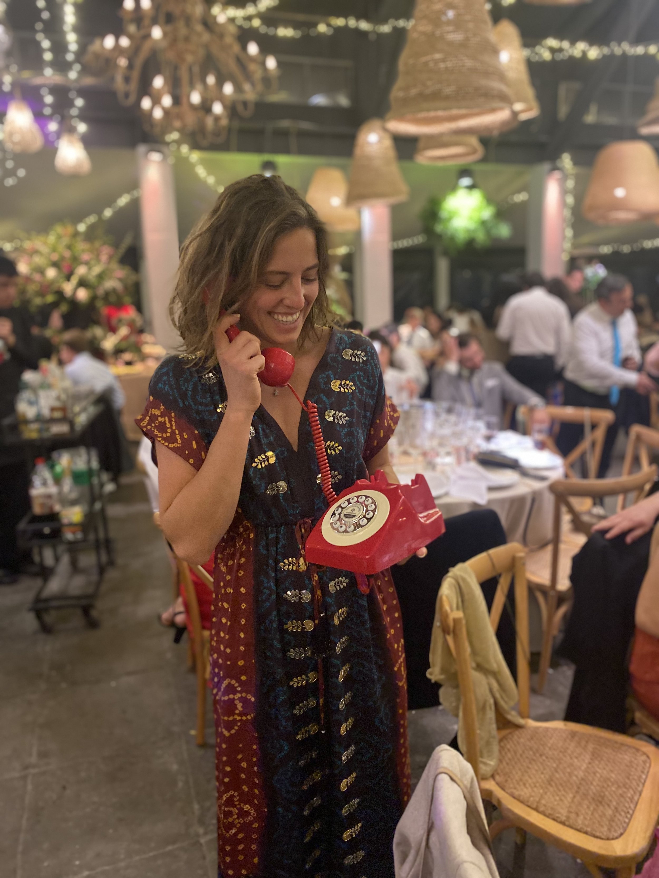Mujer en vestido colorido con sonrisa, hablando por teléfono rojo antiguo en un restaurante elegante con decoraciones de paraguas y luces, rodeada de gente.