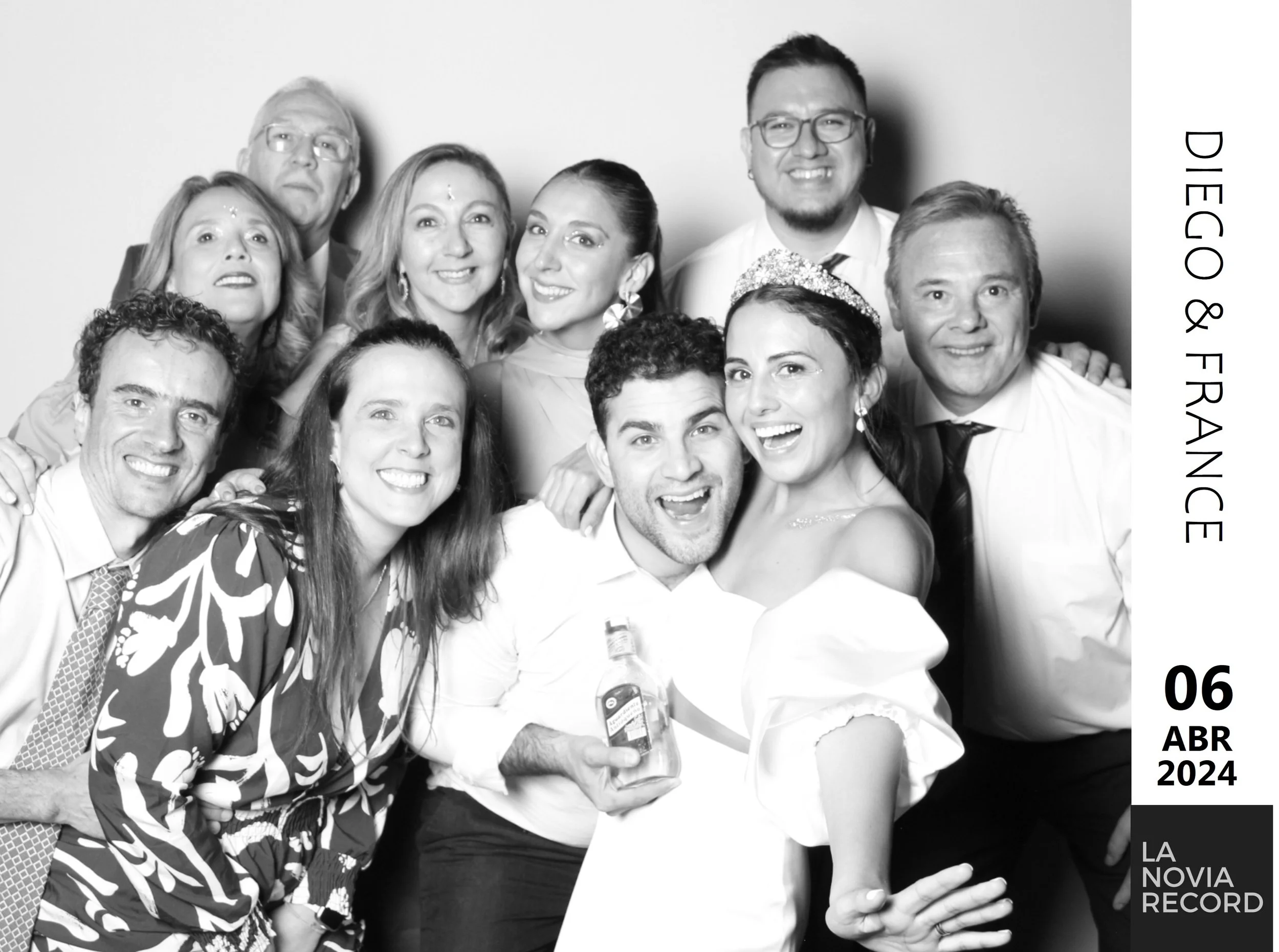 Grupo de personas en una celebración, algunos con ropa formal y accesorios, sonriendo hacia la cámara, en un fondo blanco. Fecha y evento en la esquina inferior derecha.