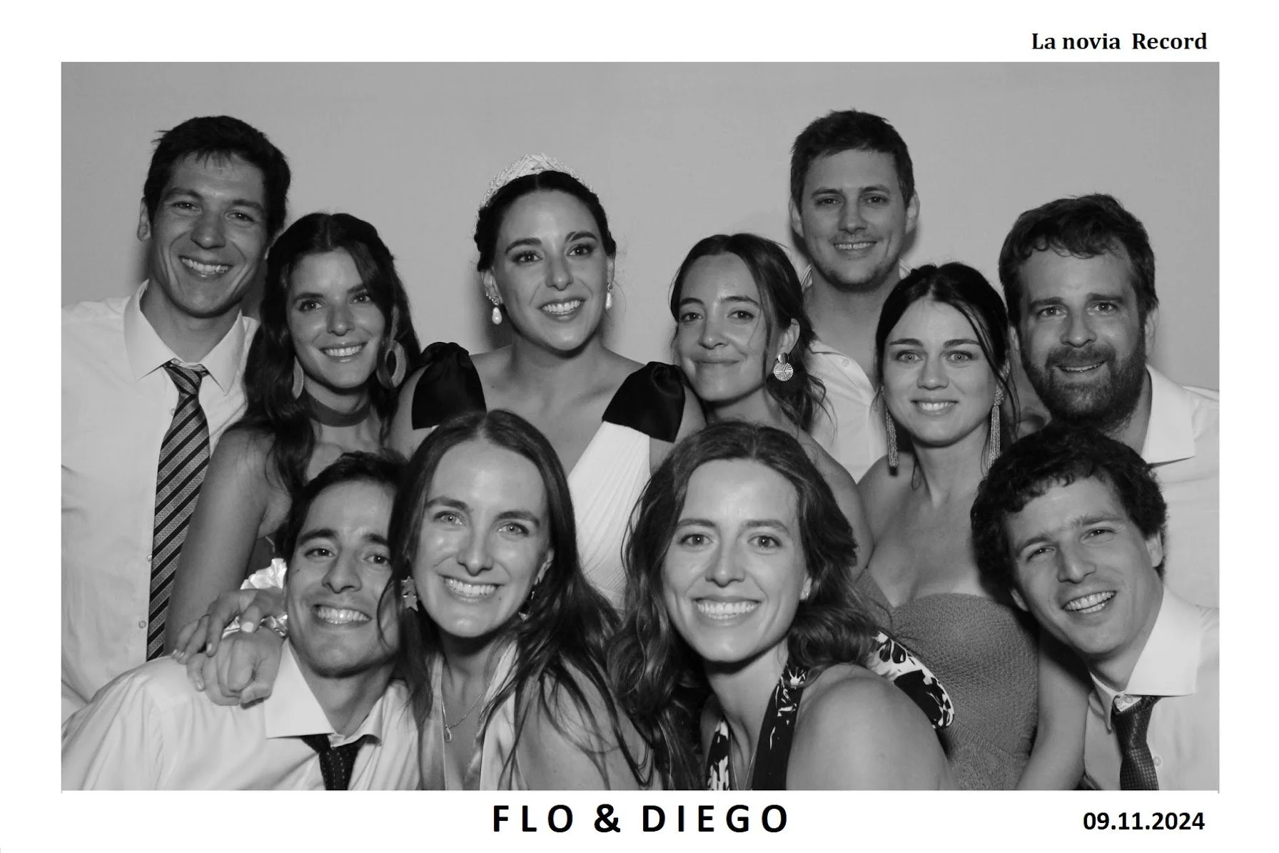 Foto en blanco y negro de un grupo de diez personas sonriendo, incluyendo una novia con diadema y vestido de novia, y un novio con corbata, en una celebración de boda. En la esquina superior derecha dice 'La novia Record', en la parte inferior están 