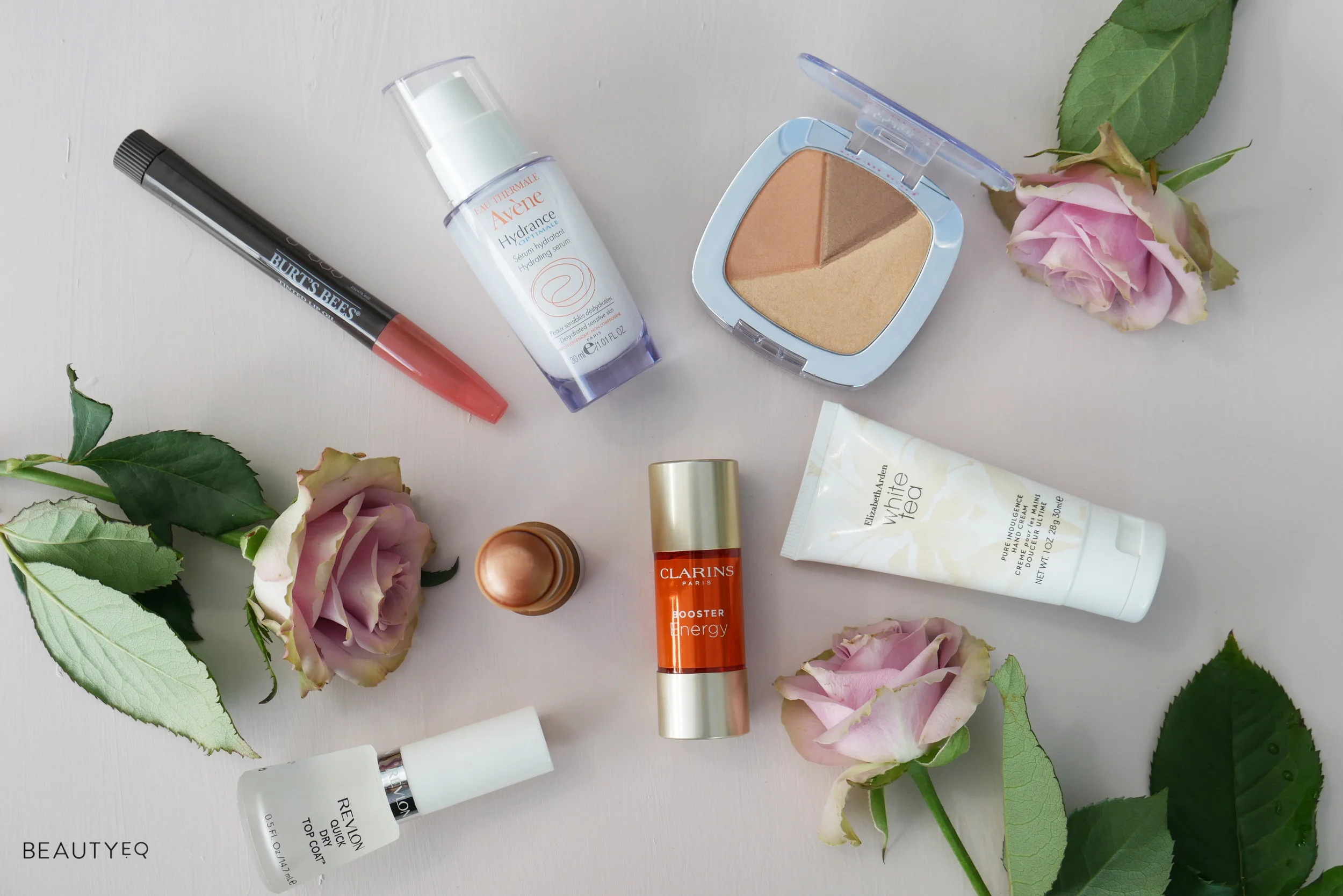 A selection of different beauty products arranged against a white wall with pink roses surrounding them 