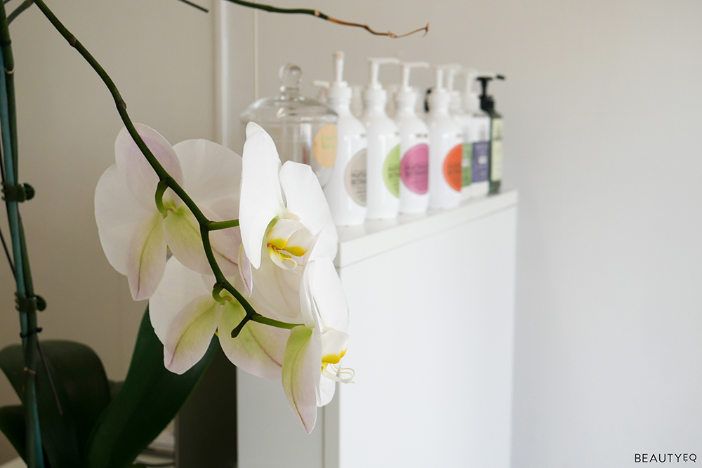 A range of beauty products in white bottles sitting on a white bench and white flowers in the corner