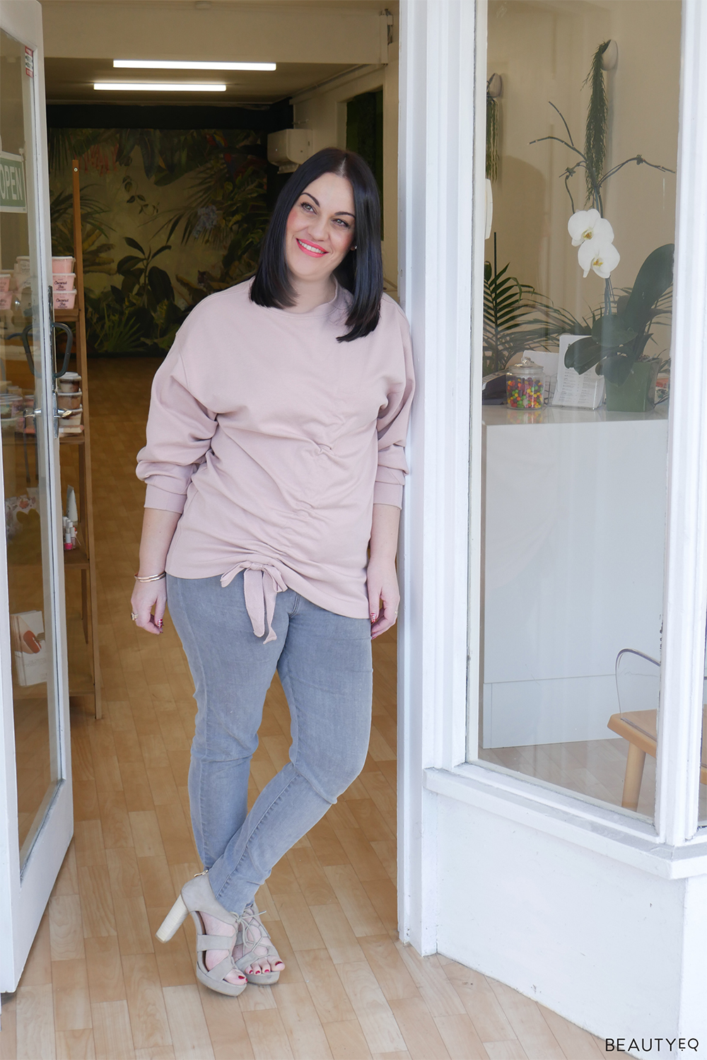 dark haired woman in blush sweater and grey jeans leaning against the white entrance way of a salon